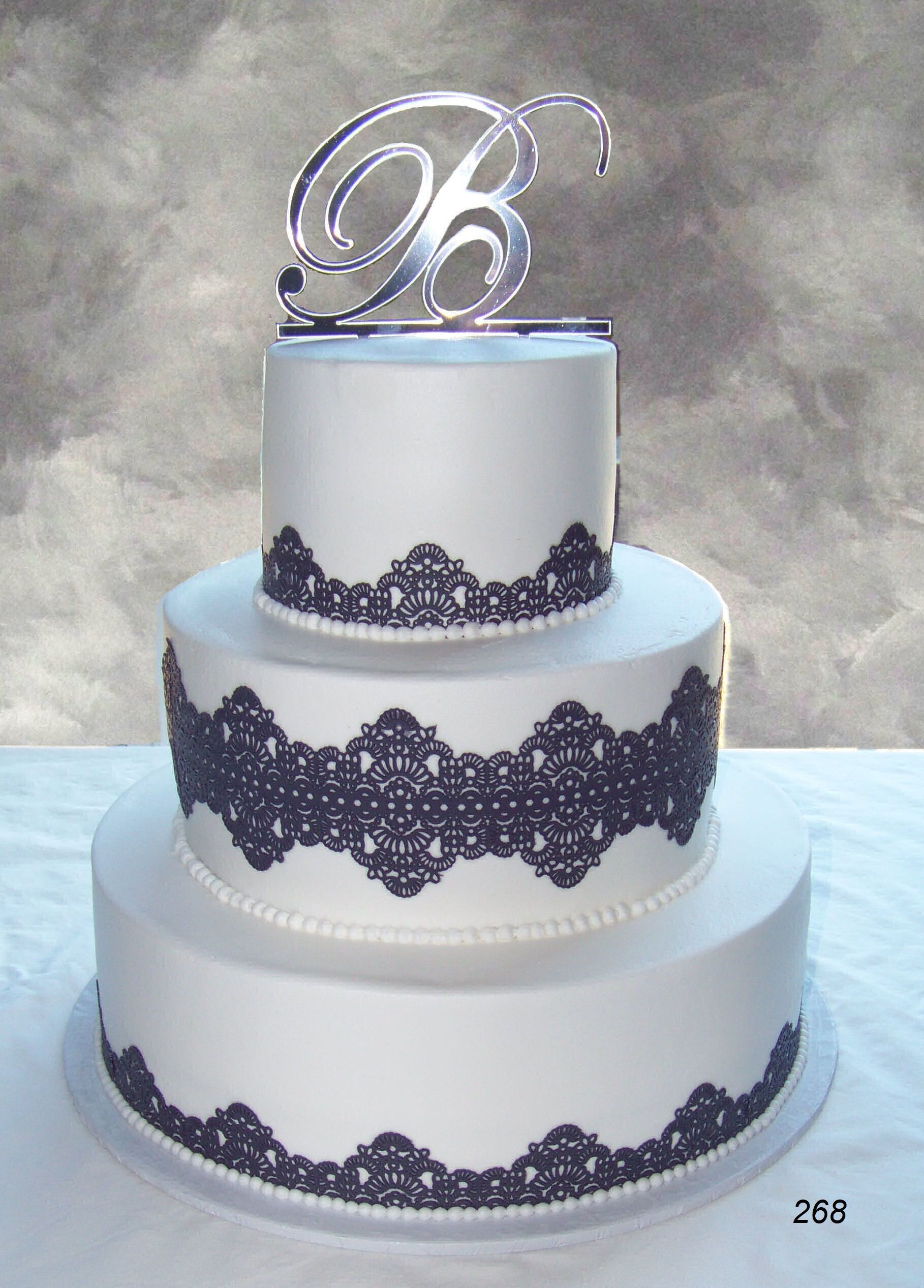 Three-tier white wedding cake with dark lace trim and a silver monogram topper