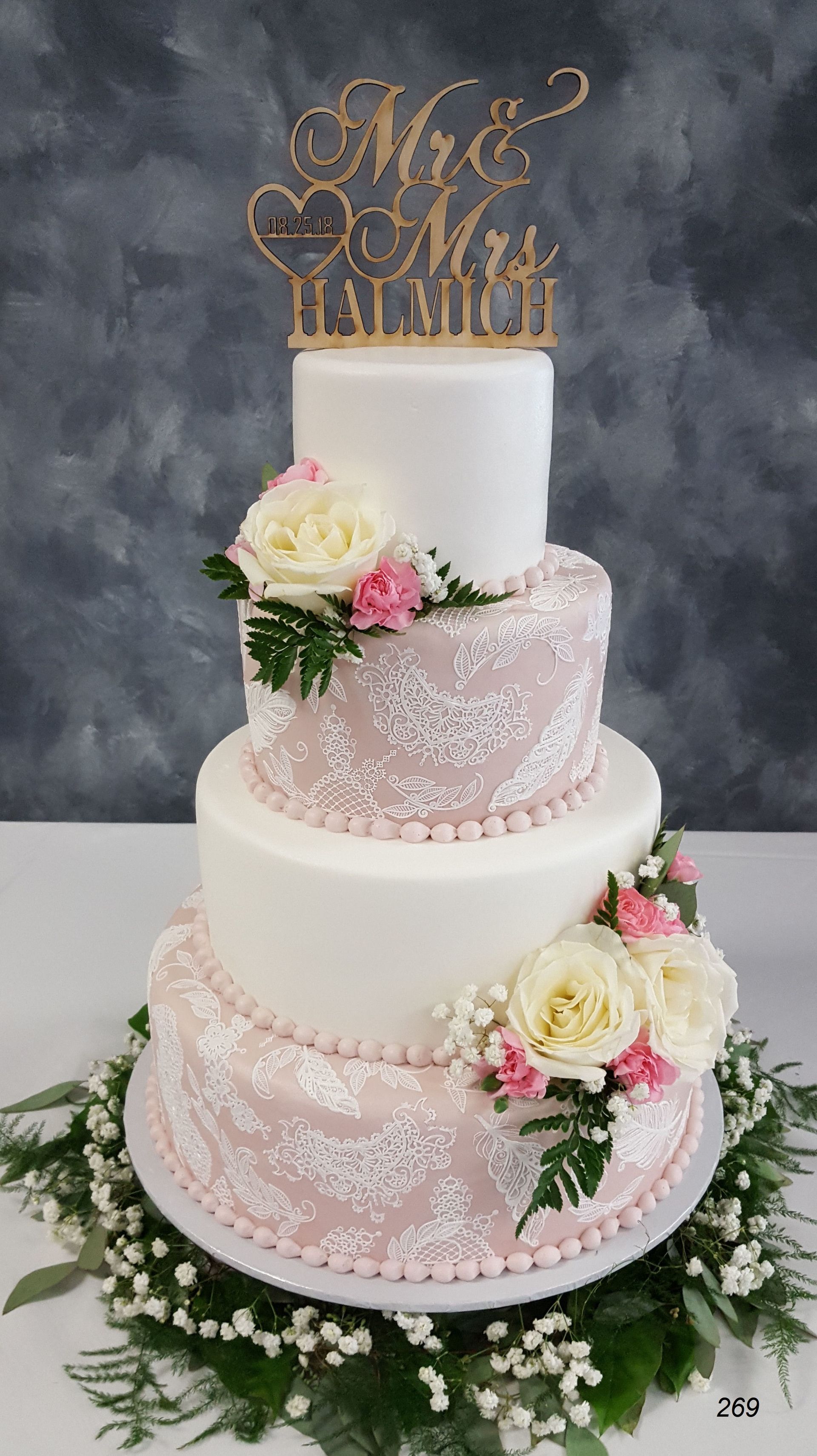Three-tier white wedding cake with blush lace, pink roses, and gold topper on a floral base