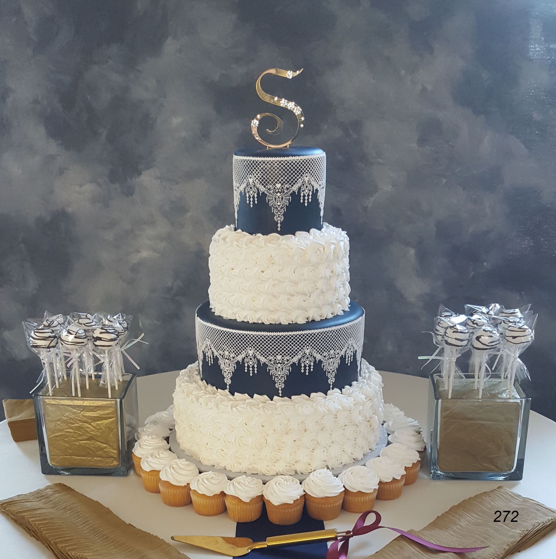Three-tier navy and white cake with gold topper, flanked by gift boxes and cupcakes on a table.
