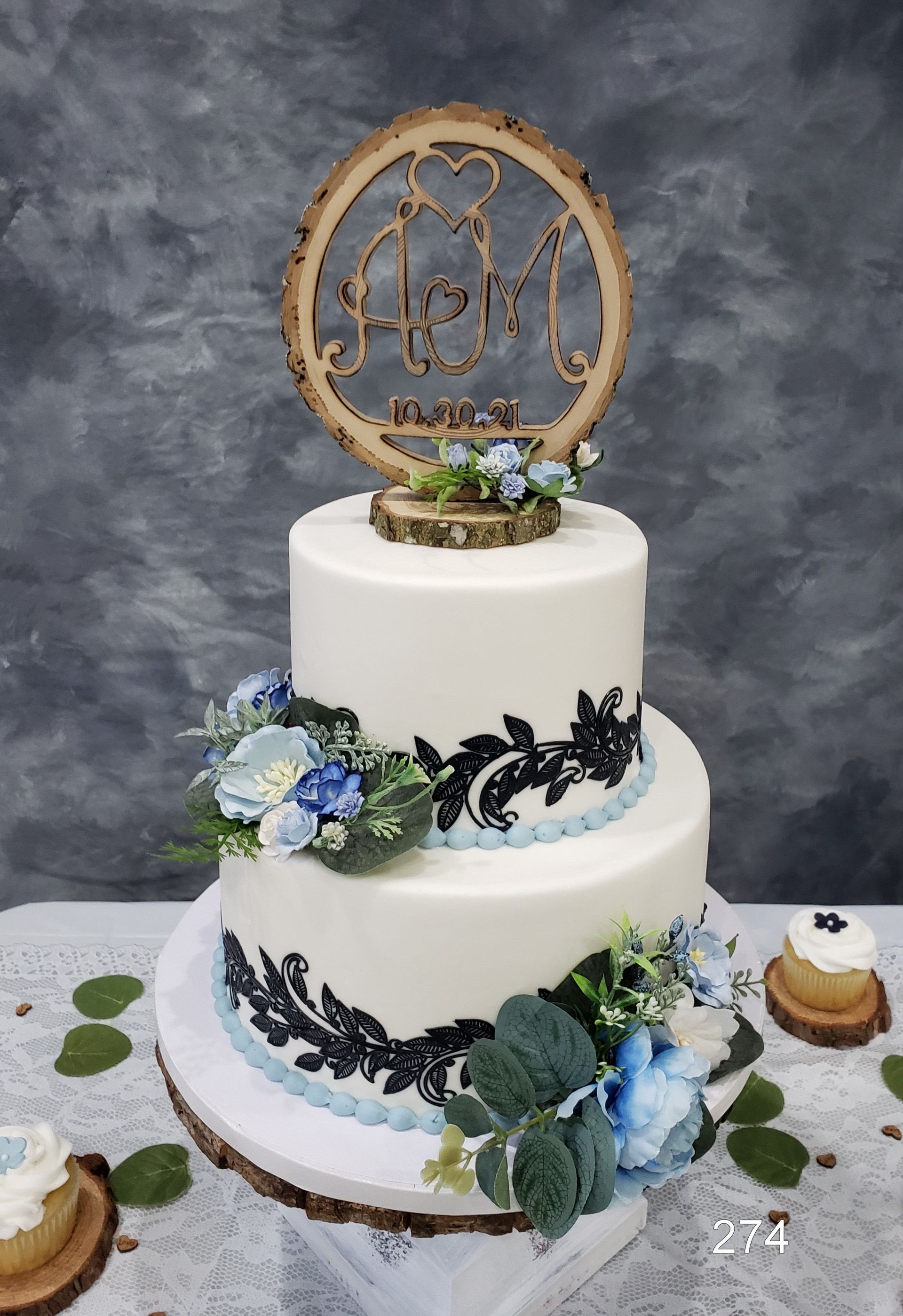 Three-tier white cake with blue flowers, black lace accents, and a gold monogram topper