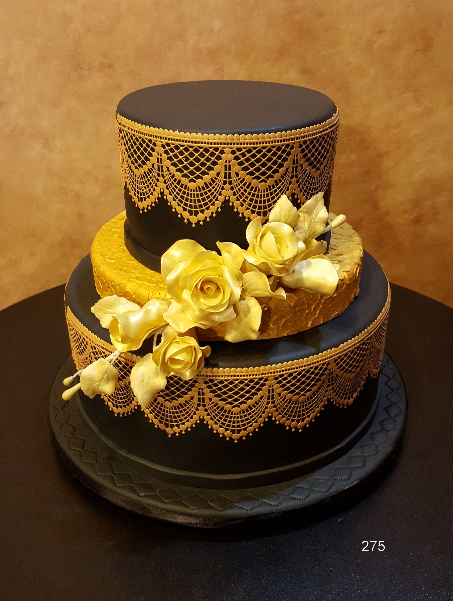 Two-tier black and gold cake with lace-like trim and yellow roses on a dark stand