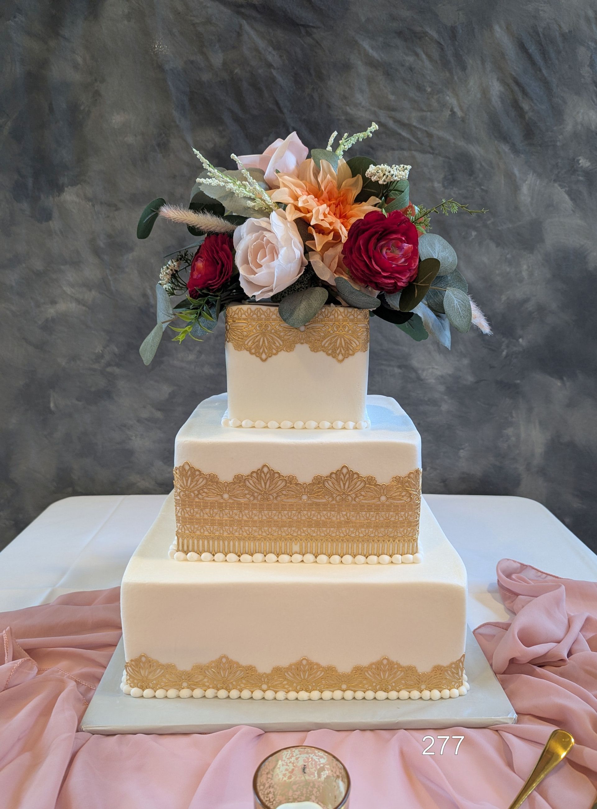 Three-tier white cake with gold accents and pink-red floral topper on a table