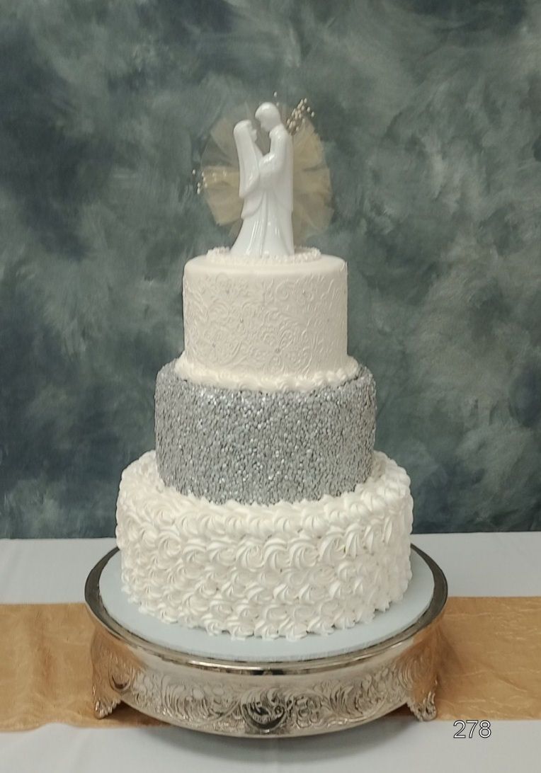Four-tier white and silver wedding cake with a bride and groom topper on a decorative stand.