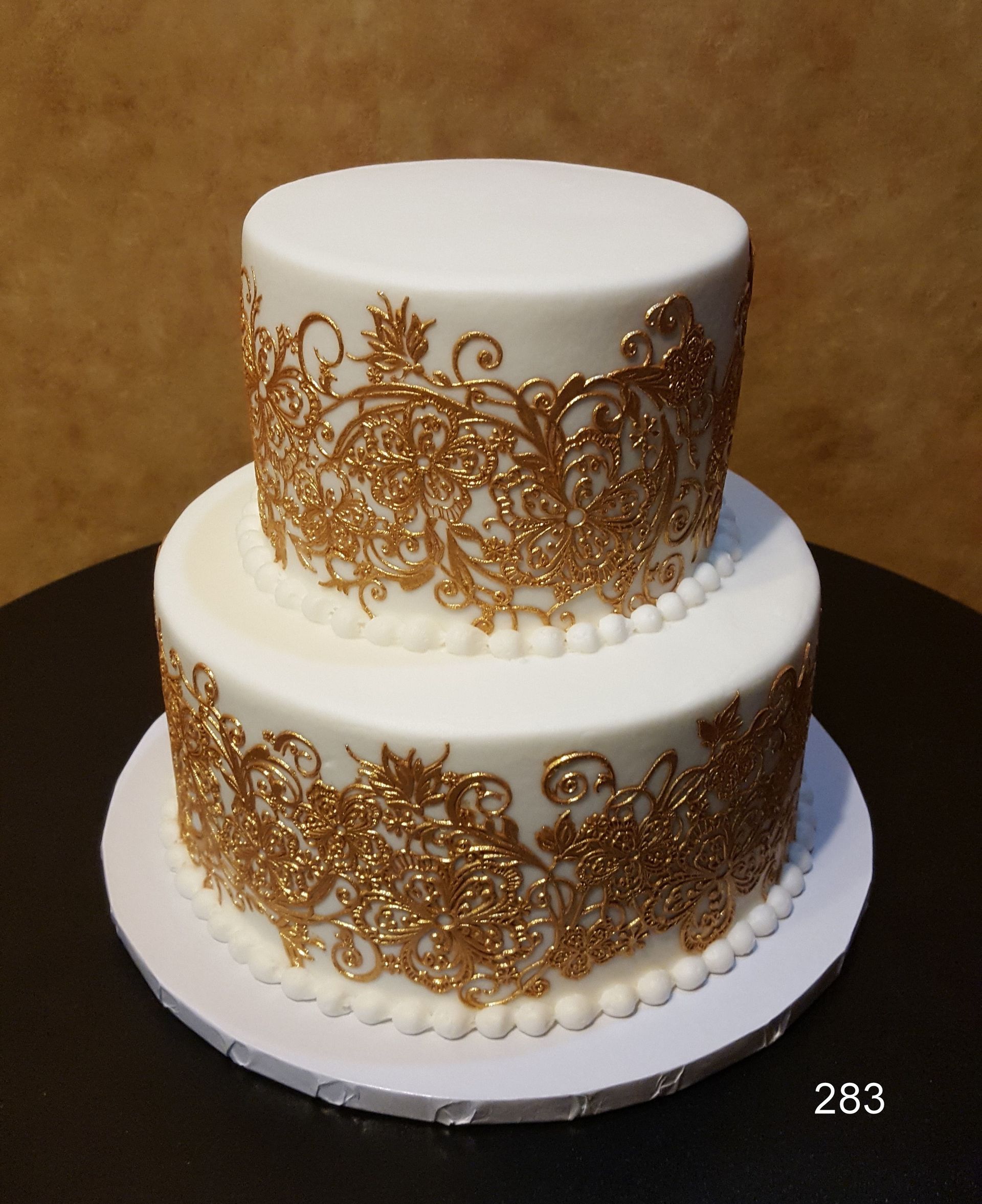 Two-tier white cake with ornate gold scrollwork and pearl borders on a white stand