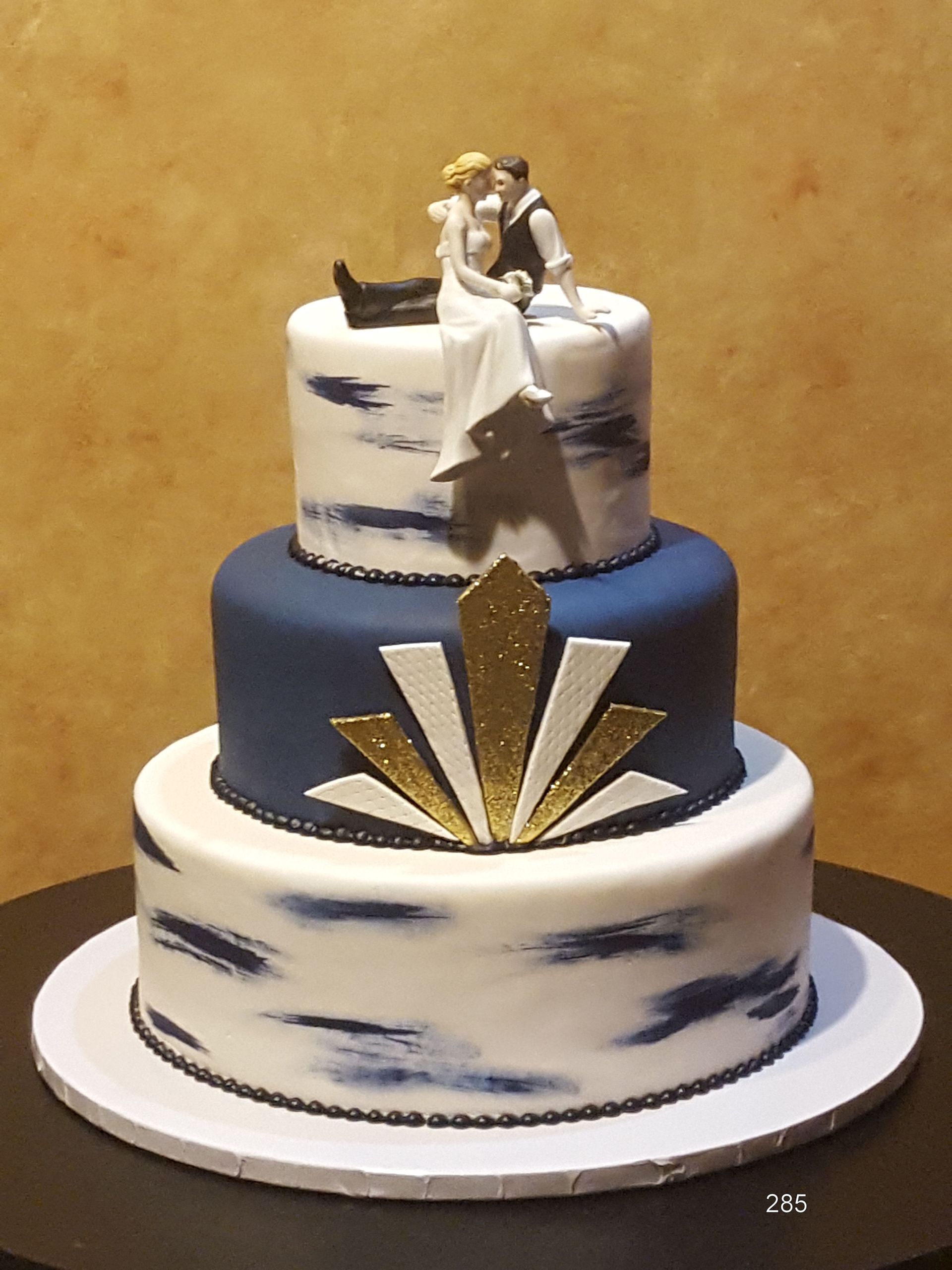 Three-tier navy, white, and gold wedding cake with a bride and groom topper and geometric gold accents