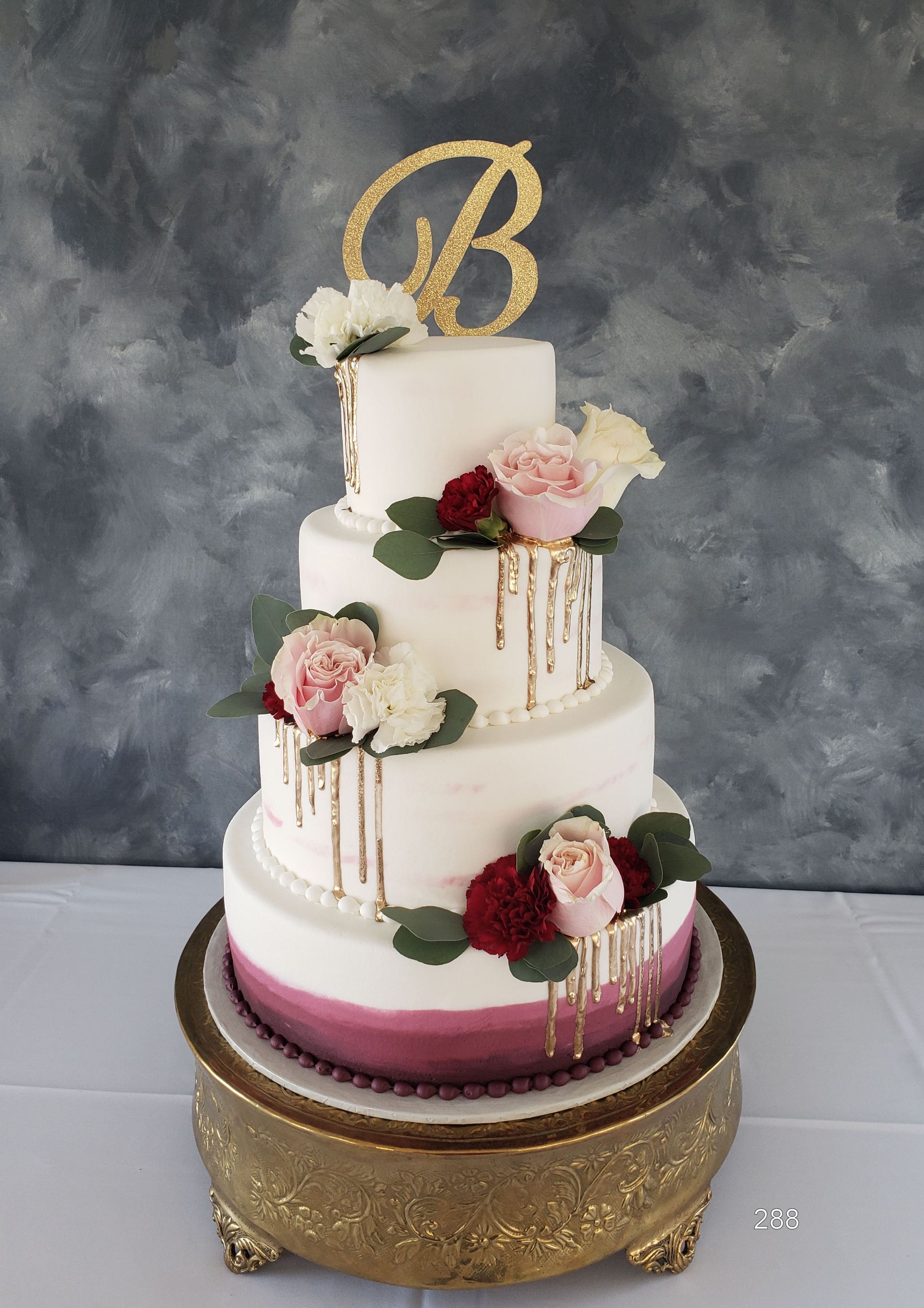 Three-tier white and blush wedding cake with gold drips, pink flowers, and gold monogram topper