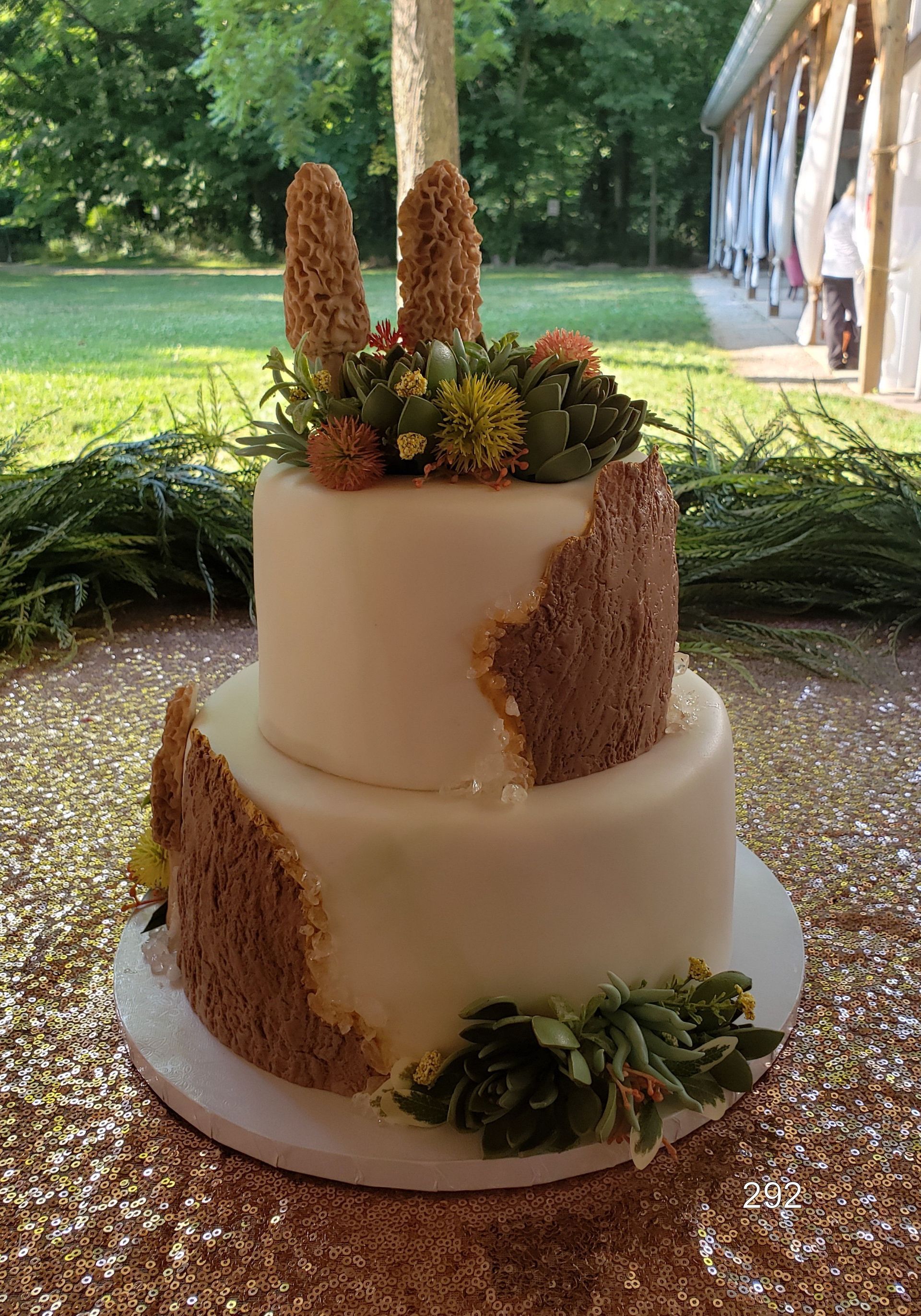 Two-tier white wedding cake with greenery and gold accents on a rustic table outdoors