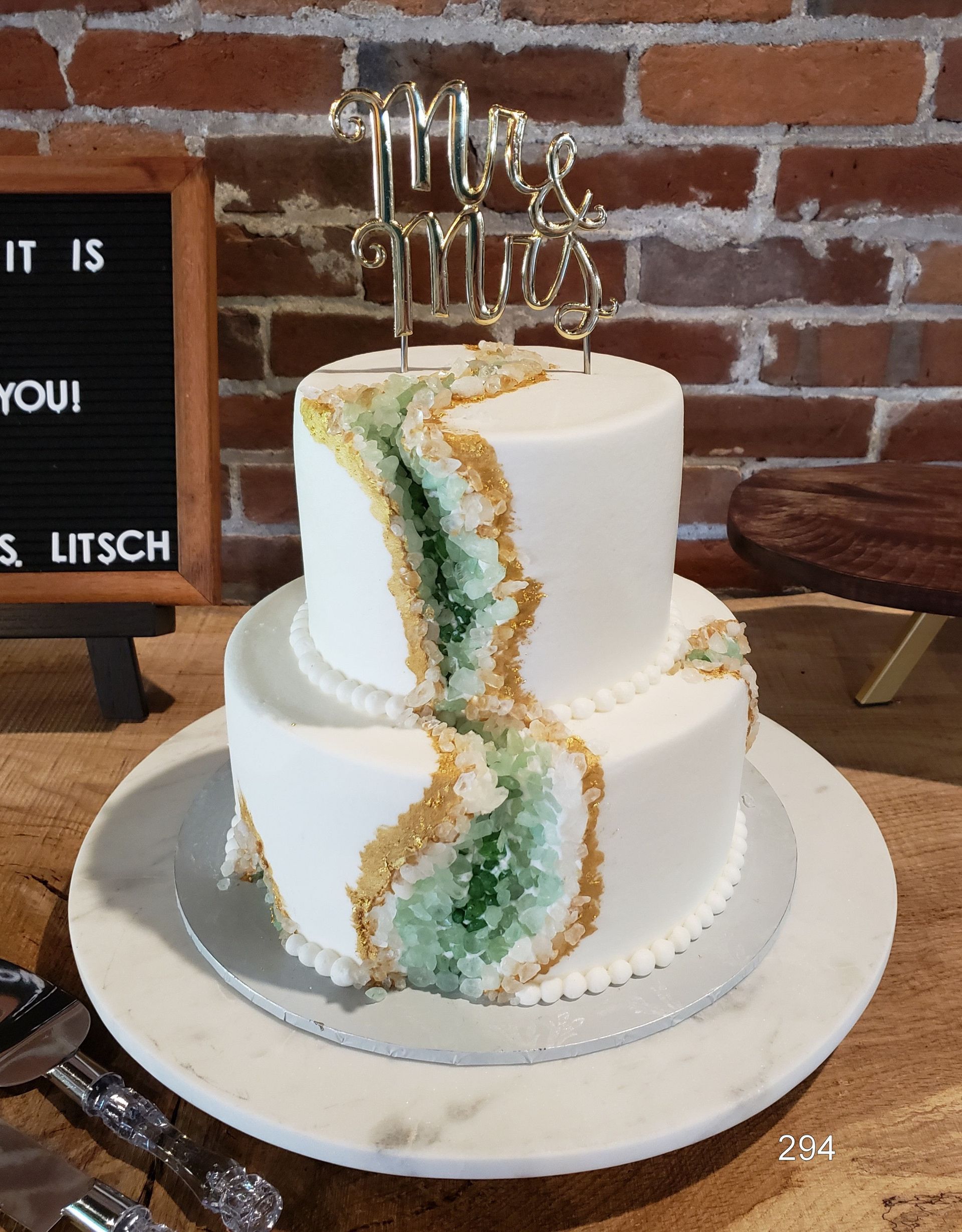 Two-tier white cake with a green and gold geode design on a plate against a brick wall
