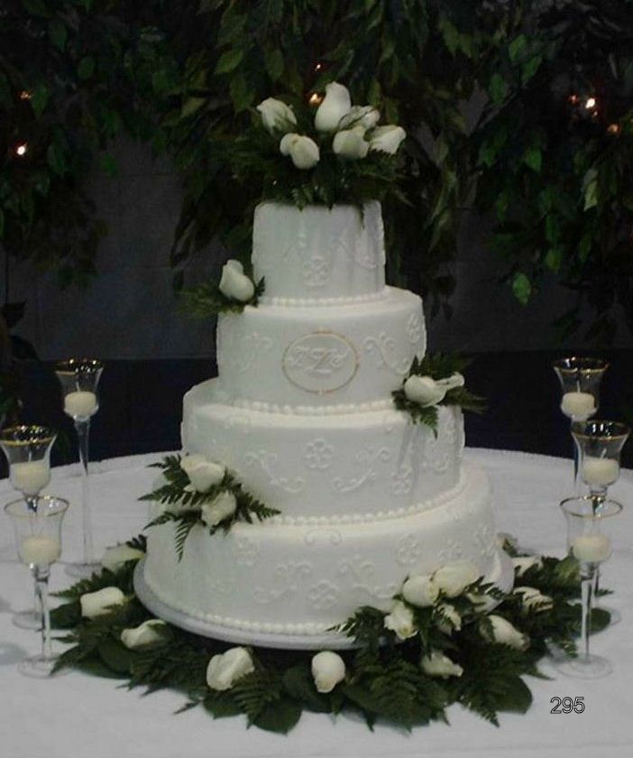 Three-tier white wedding cake with white roses and greenery on a table, surrounded by glasses