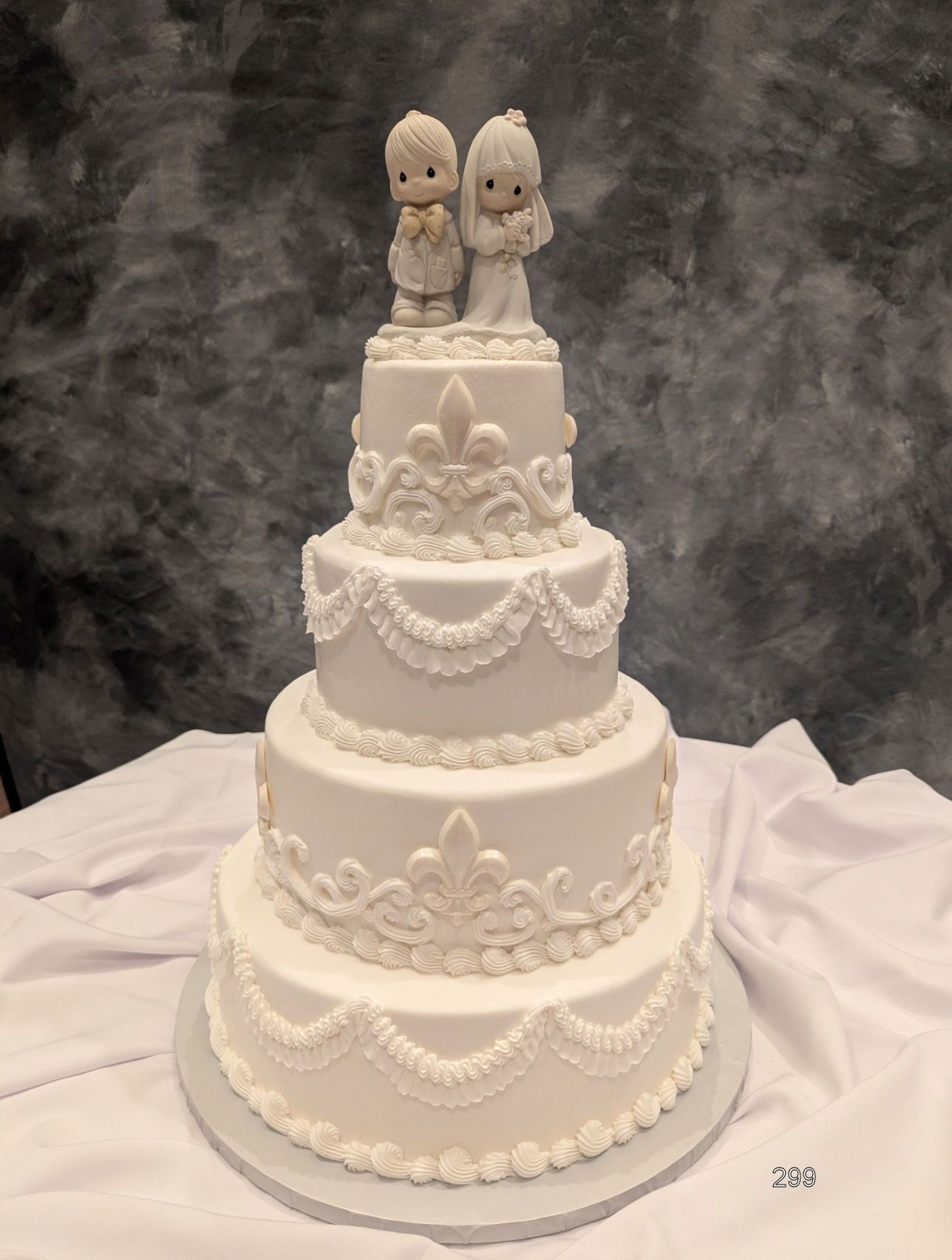 White multi-tiered wedding cake with ornate icing and two figurines on top against a dark backdrop