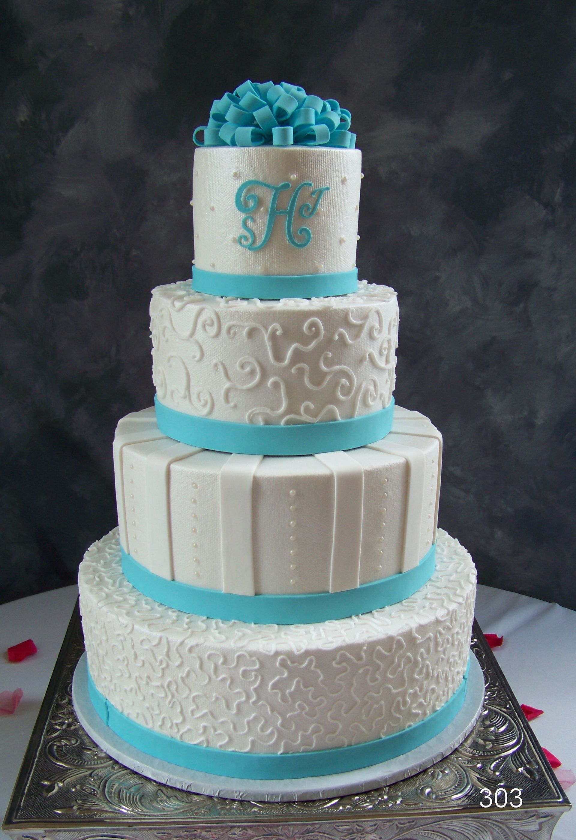 Four-tier white cake with turquoise accents and a turquoise flower topper on a silver board