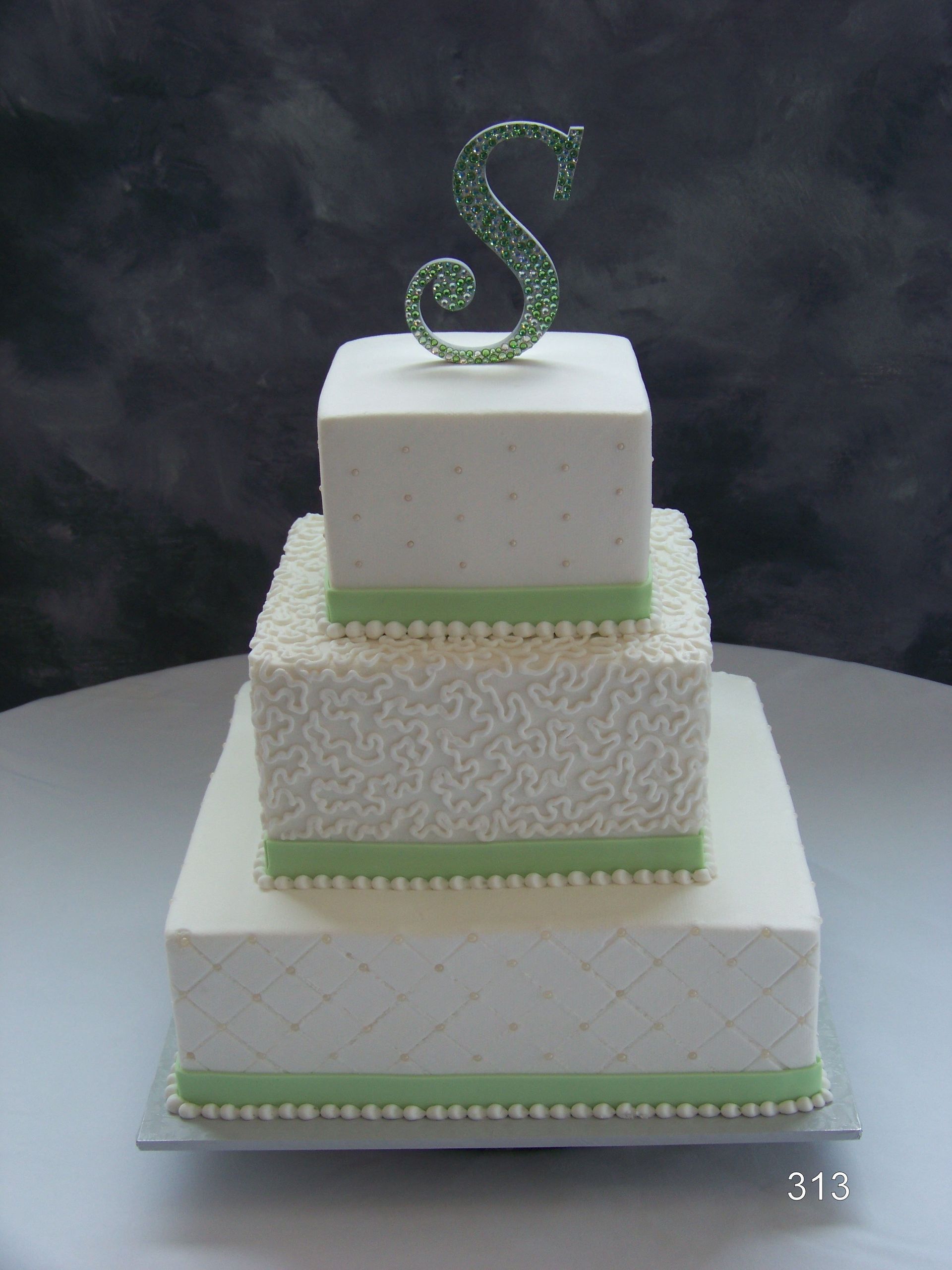 Three-tier white cake with green trim and a green jeweled letter S topper against a dark backdrop