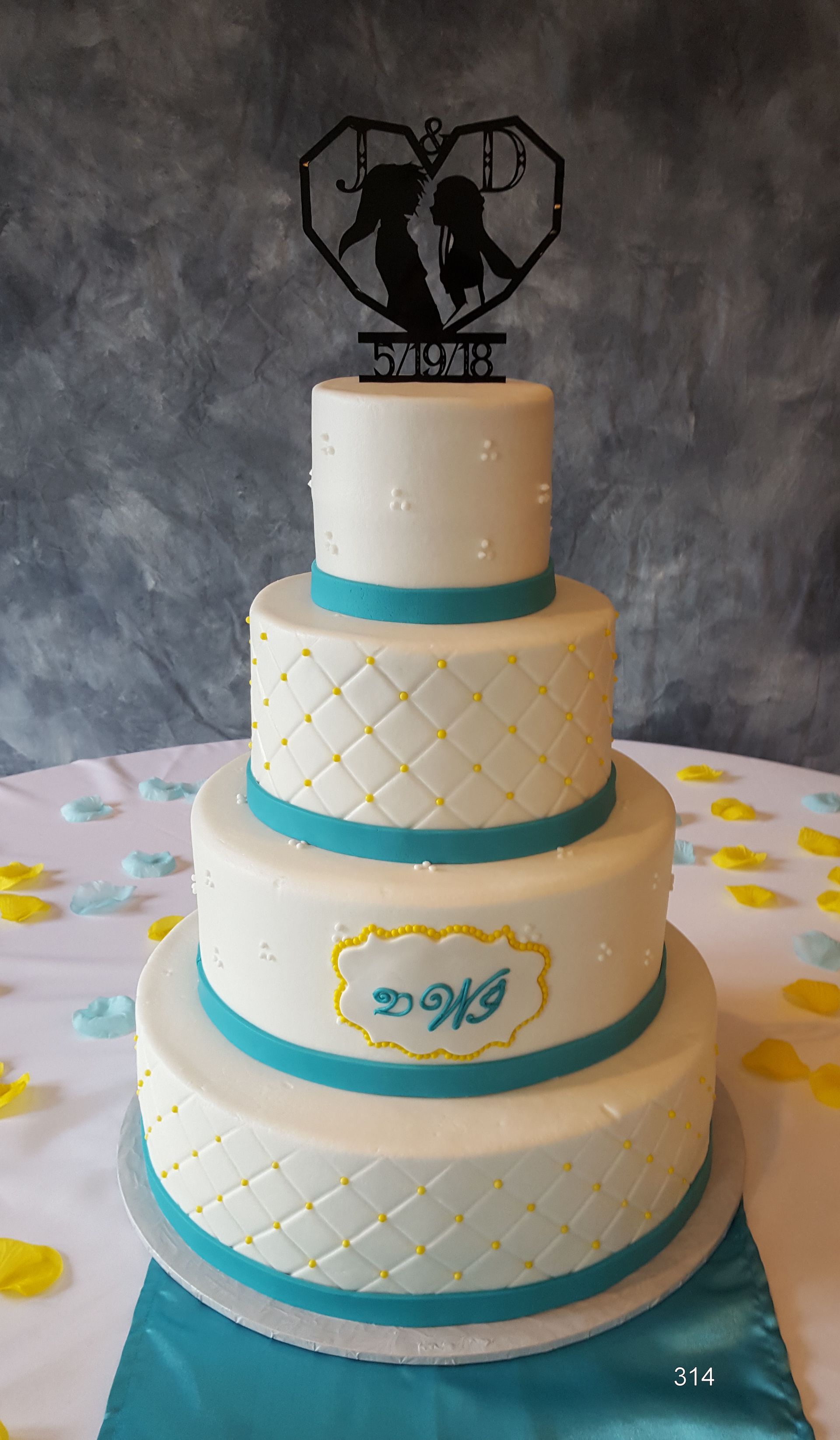 Four-tier white and gold wedding cake with turquoise accents and heart-shaped topper on a decorated table
