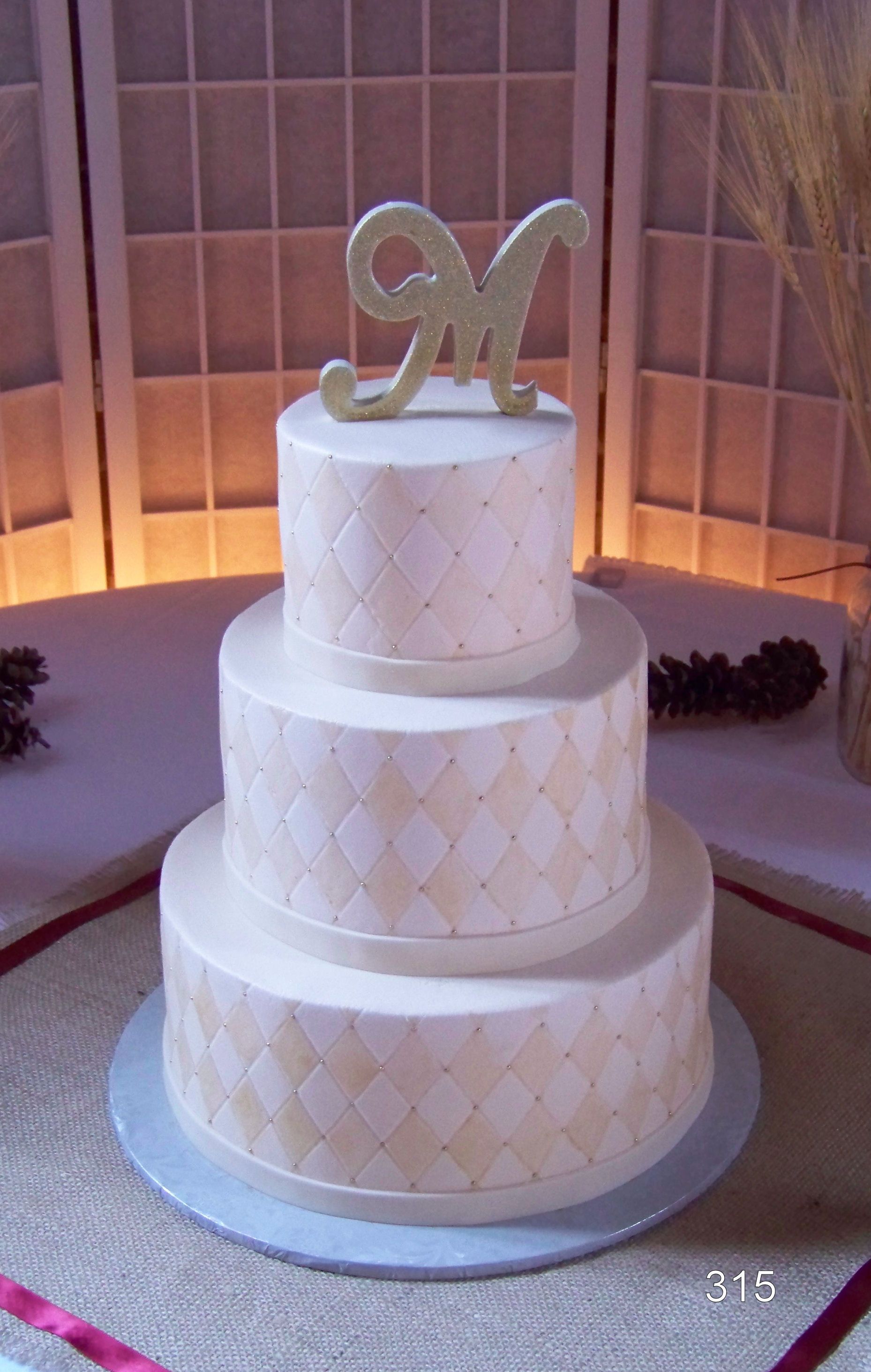 Three-tier white wedding cake with gold monogram topper in a softly lit indoor venue