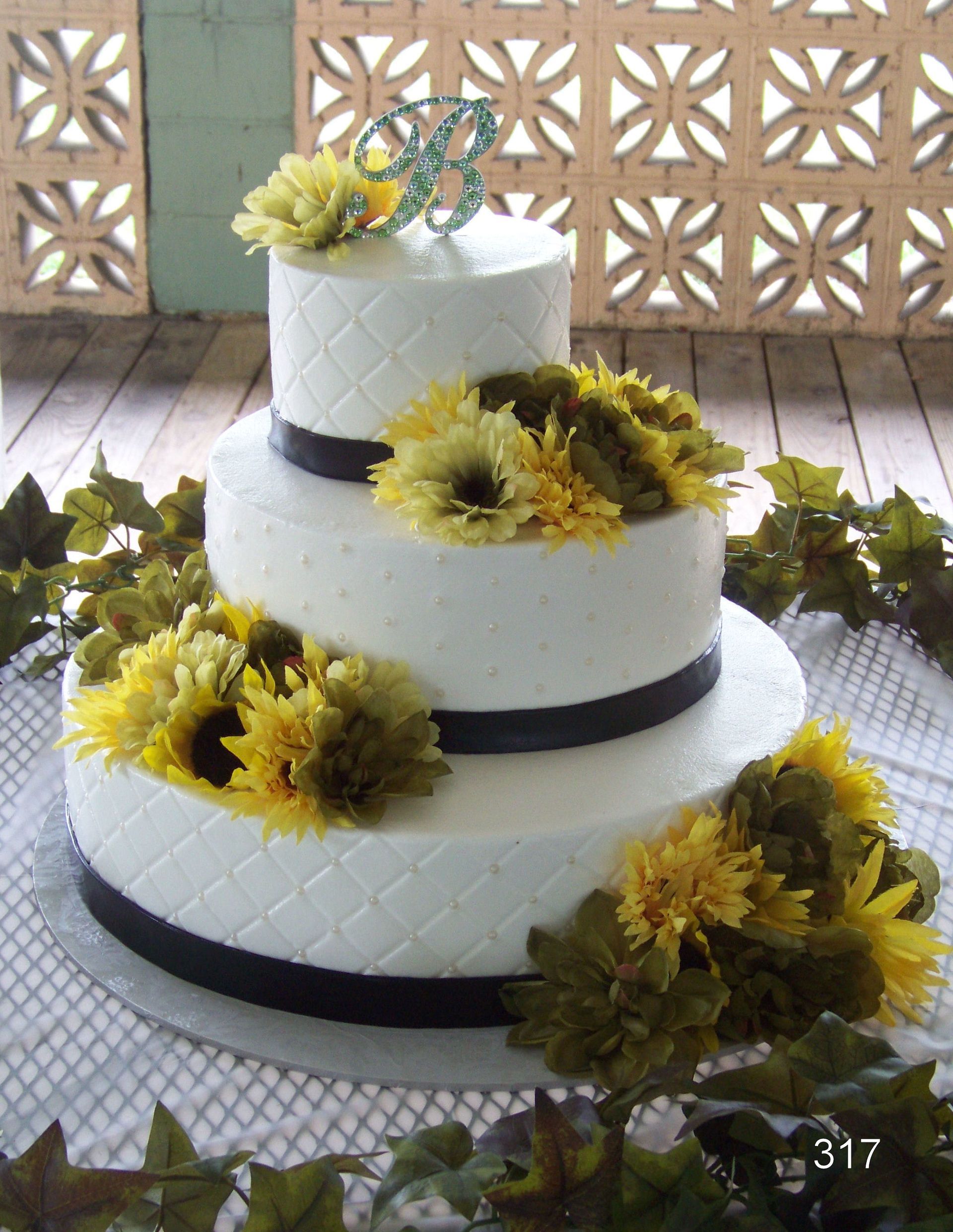 Three-tier white wedding cake with black ribbon and yellow sunflowers on a table.