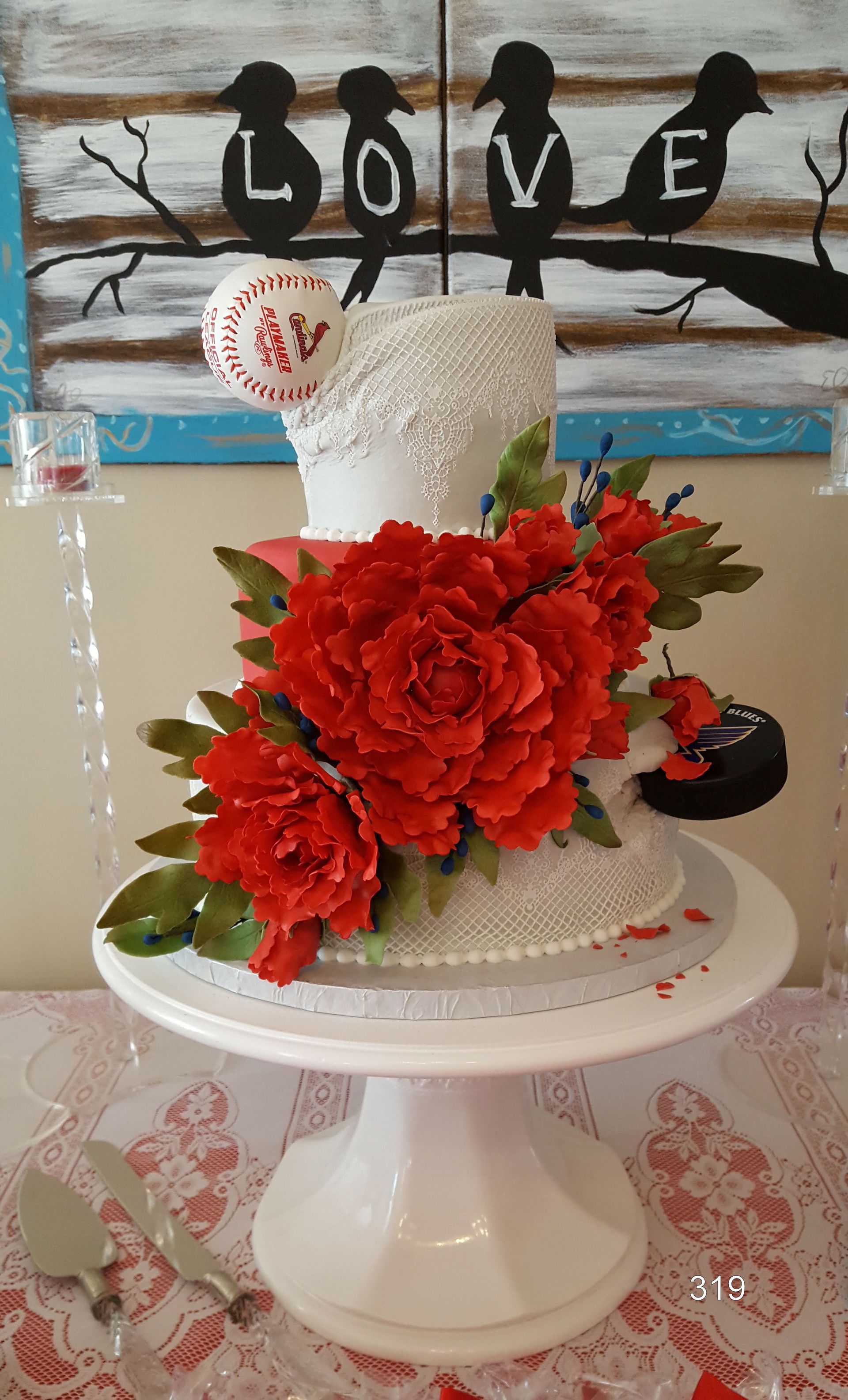 White tiered cake with bright red flowers and “LOVE” topper on a pedestal stand, outdoors.
