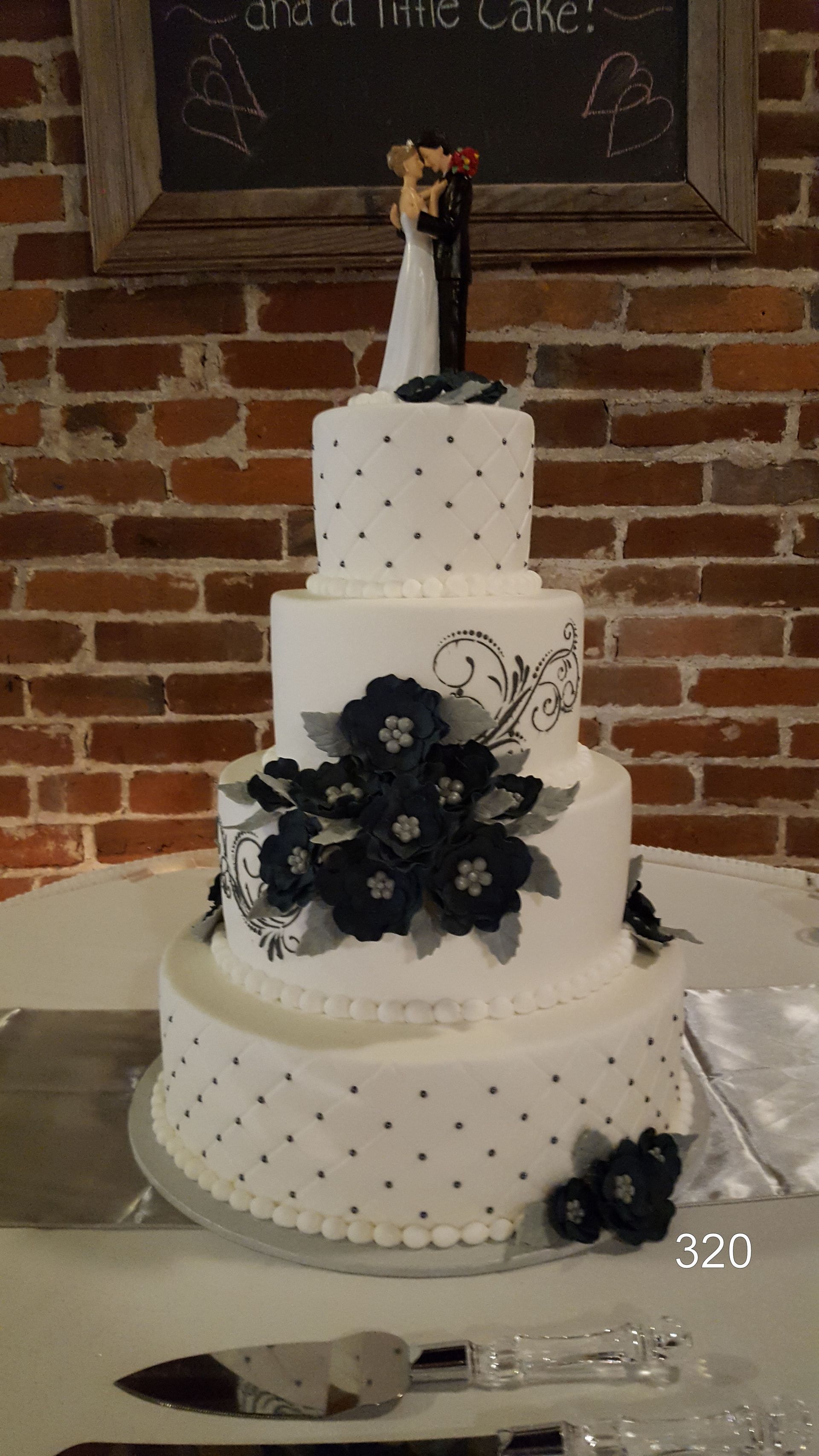 Three-tier white wedding cake with black floral decorations and a bride-and-groom topper on a brick backdrop