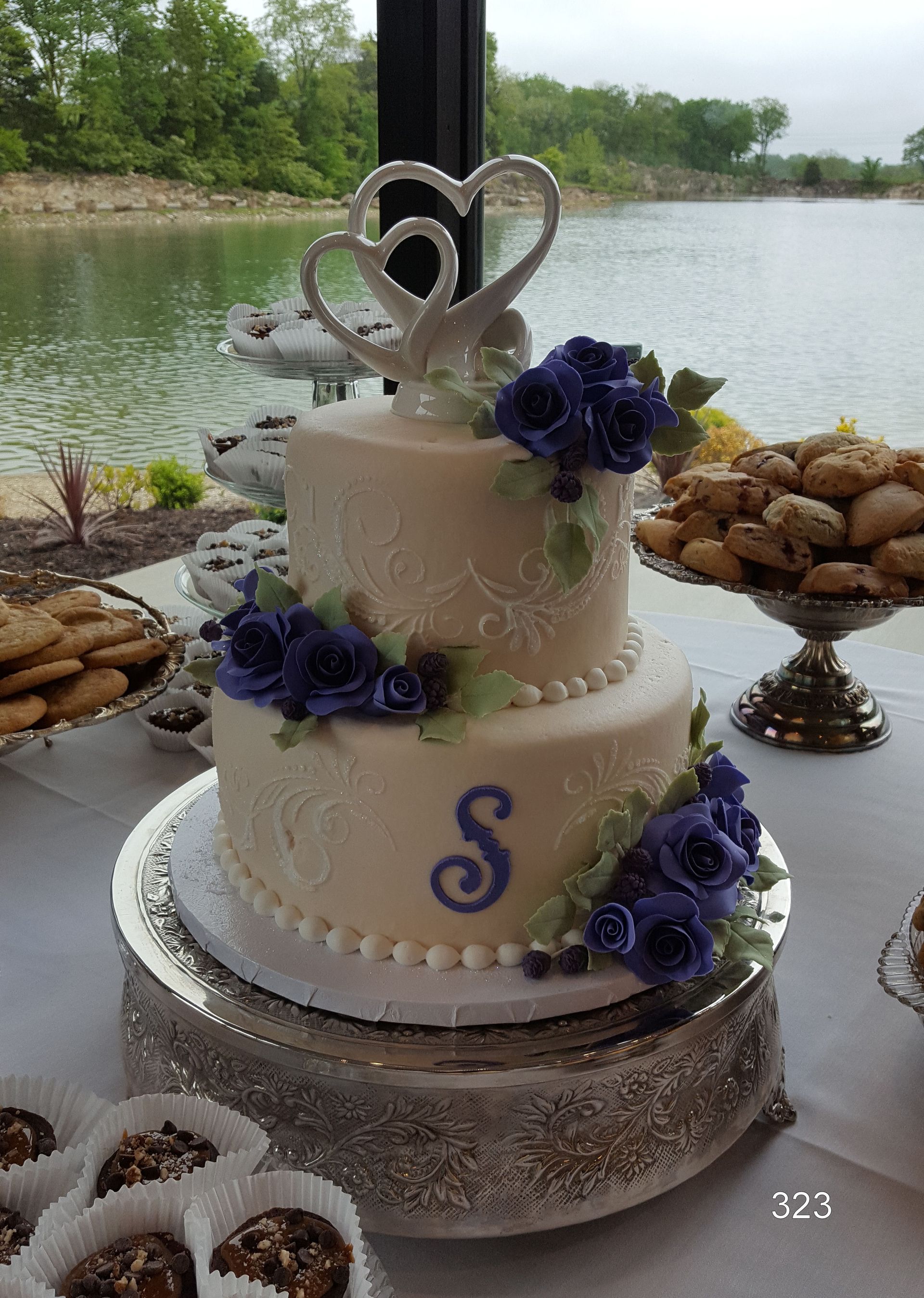 Two-tier white cake with purple flowers and heart topper on a riverside table