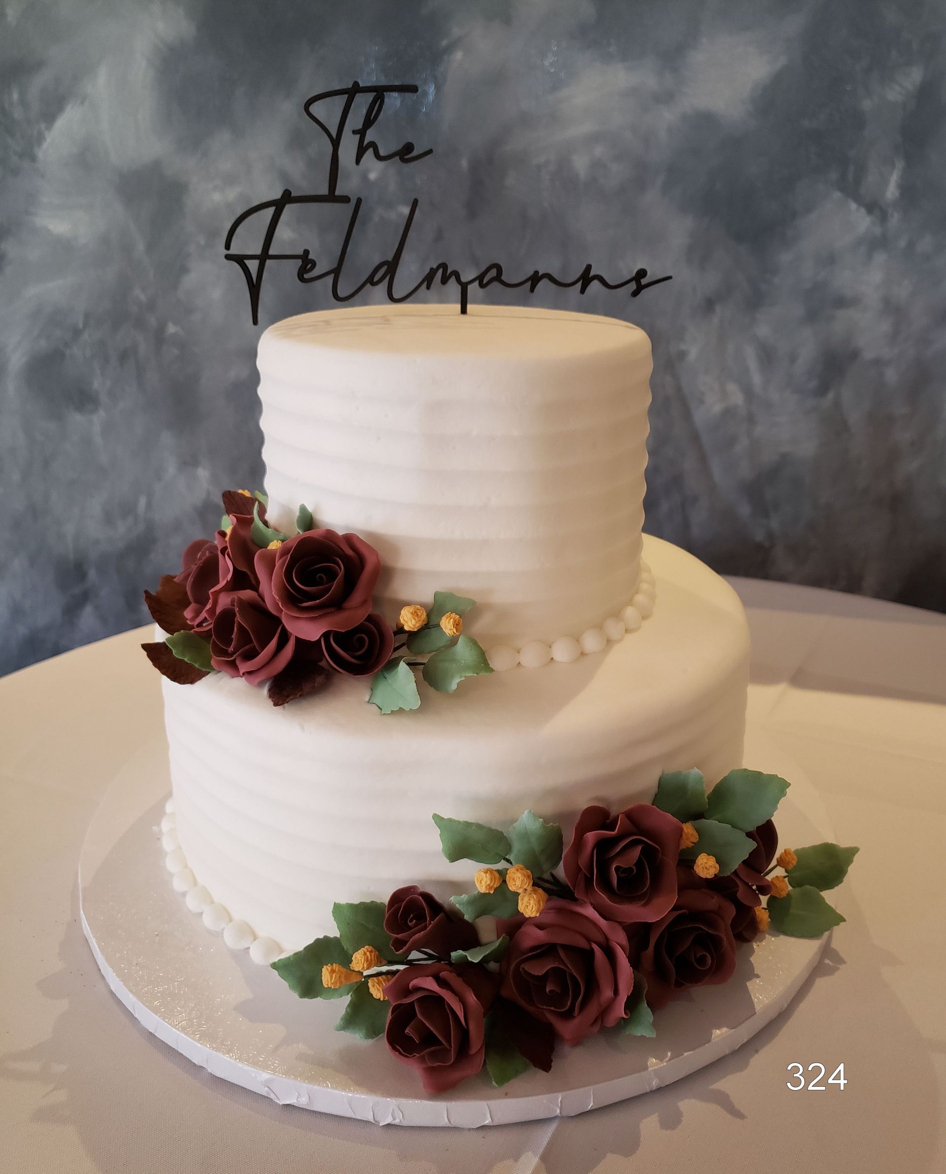 Two-tier white wedding cake with burgundy roses and greenery, topped with a scripted name topper