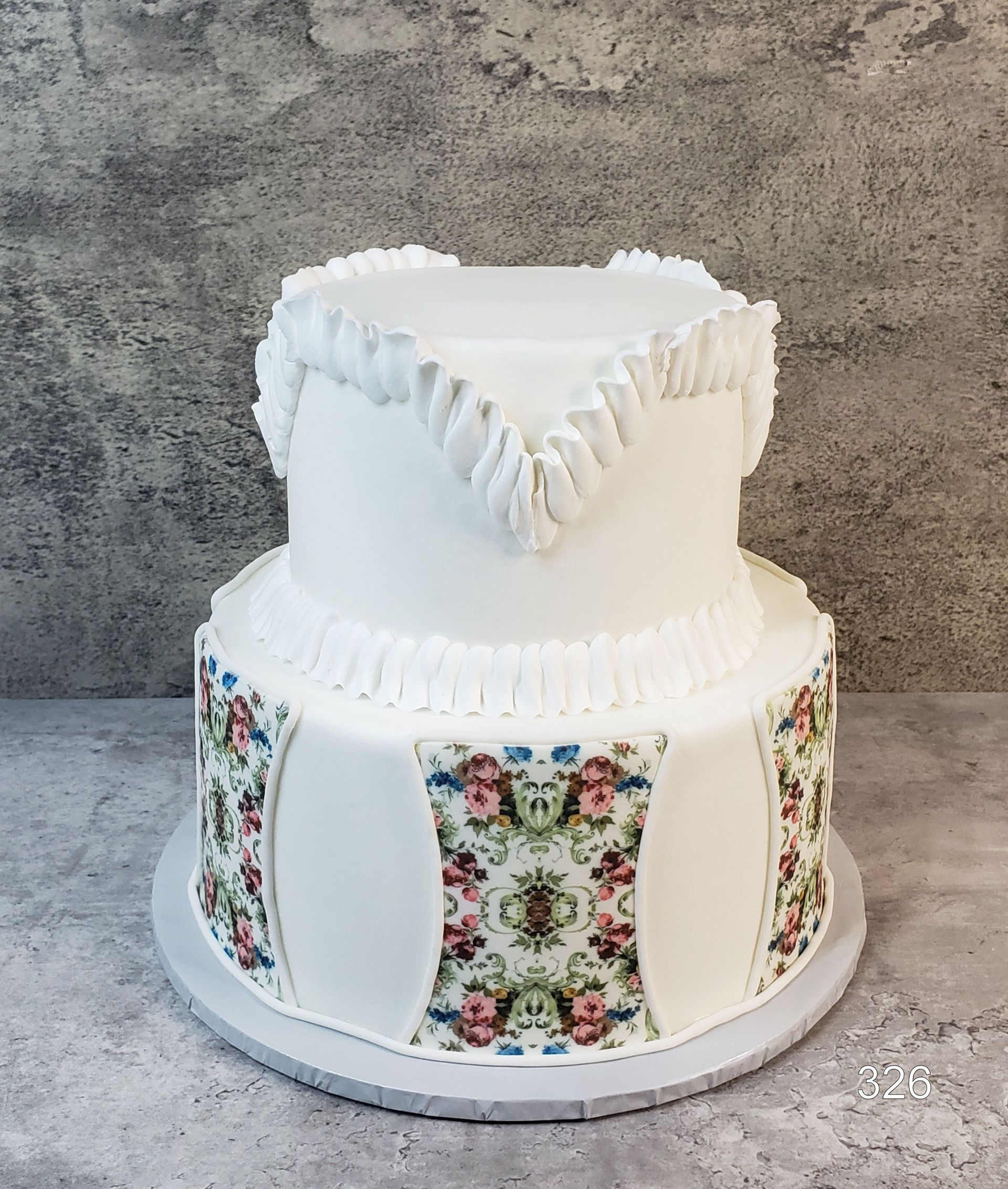 White tiered cake with floral panels and ruffled icing on a gray backdrop
