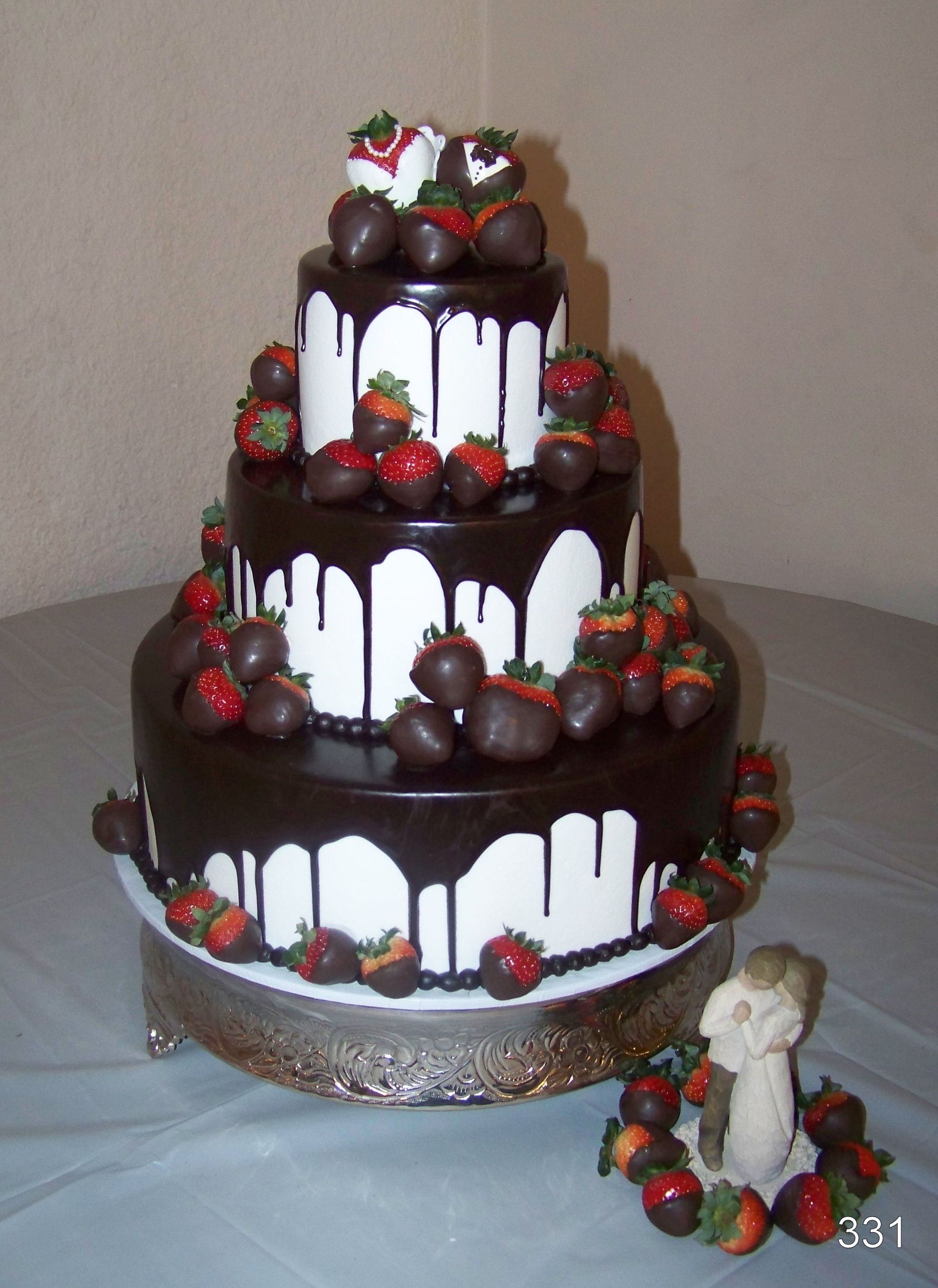 Three-tier chocolate-drip cake decorated with strawberries, on a white pedestal with an angel figurine nearby