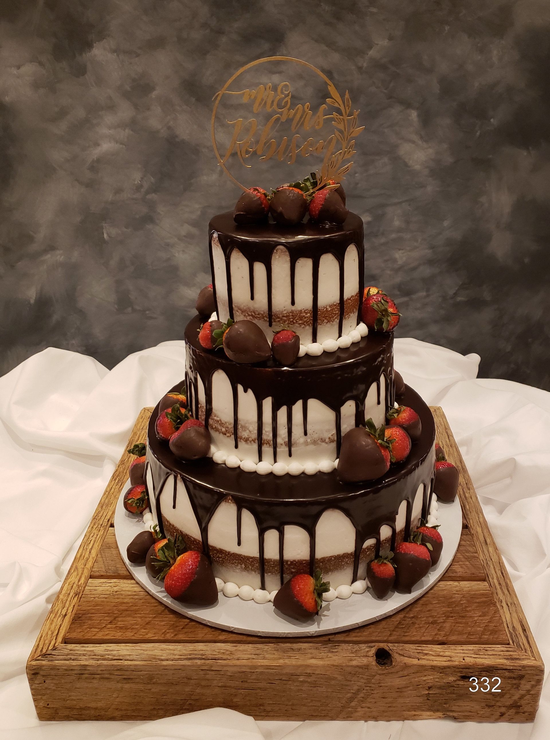 Three-tier chocolate drip cake topped with strawberries and gold decor on a wooden stand