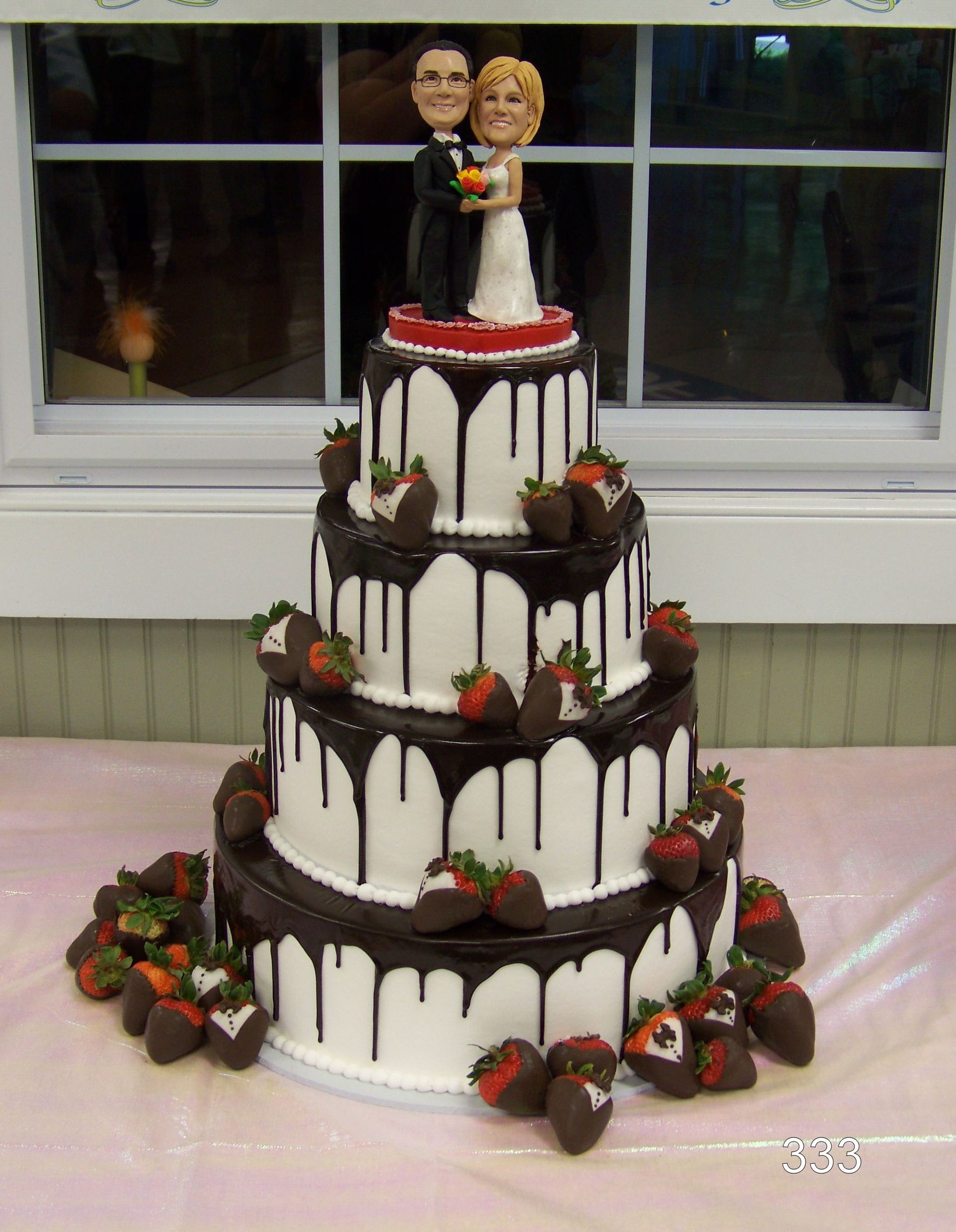 Four-tier wedding cake with chocolate drips, strawberries, and a bride-and-groom topper by a window.