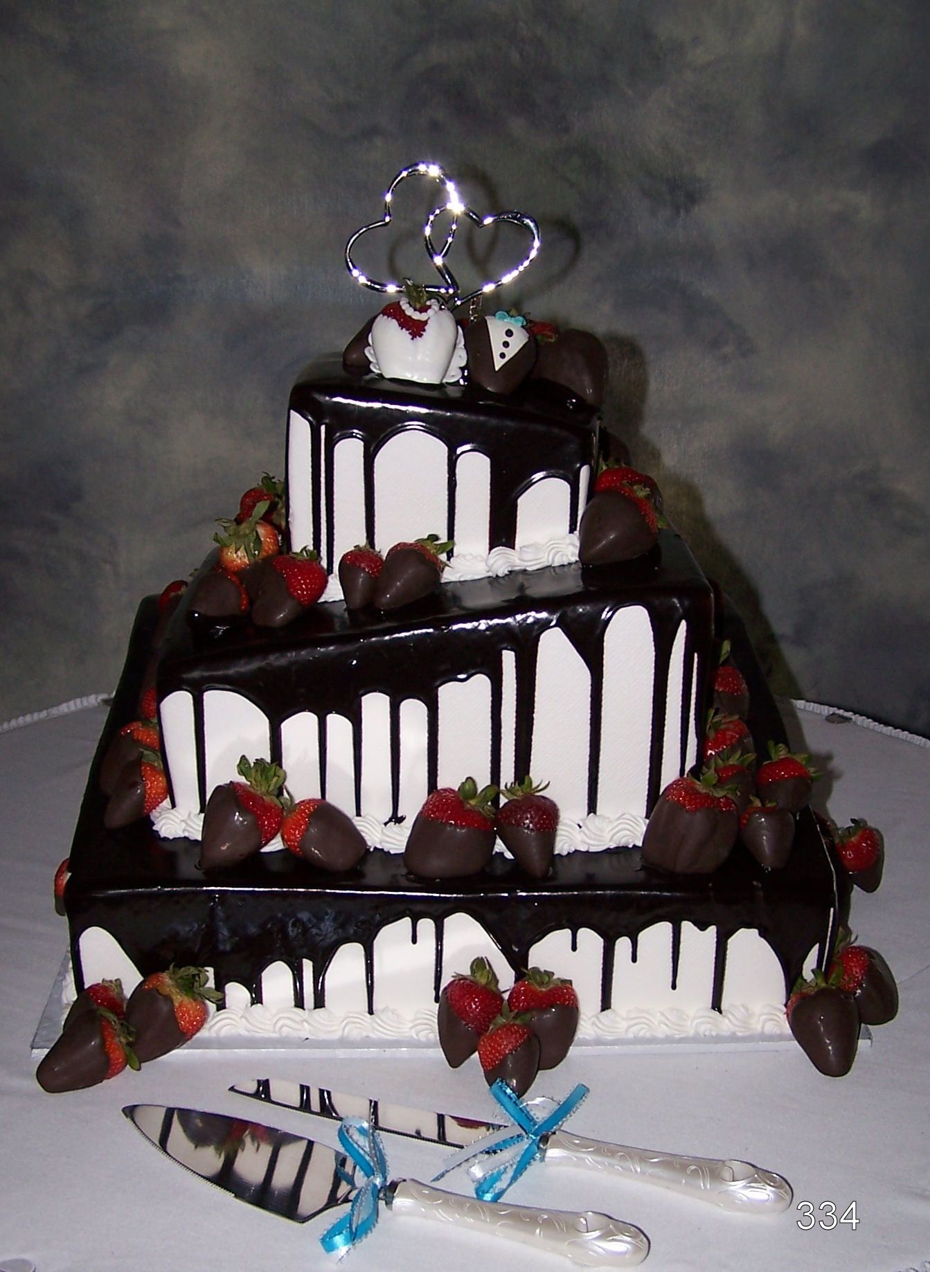 Three-tier chocolate drip cake topped with strawberries and silver swirls, on a table with ribbons.