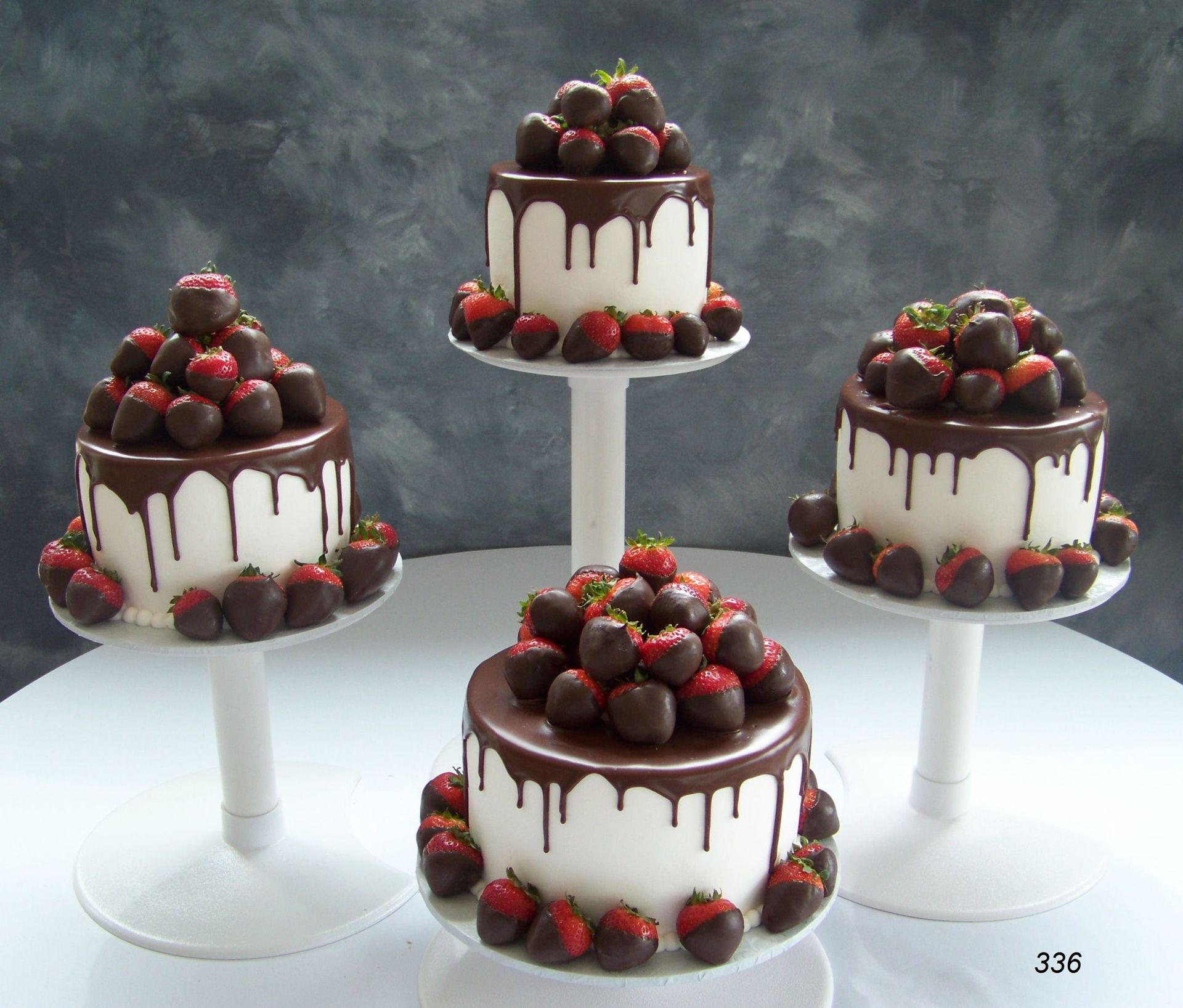 Five chocolate-drizzled strawberry cakes on white stands against a gray background.