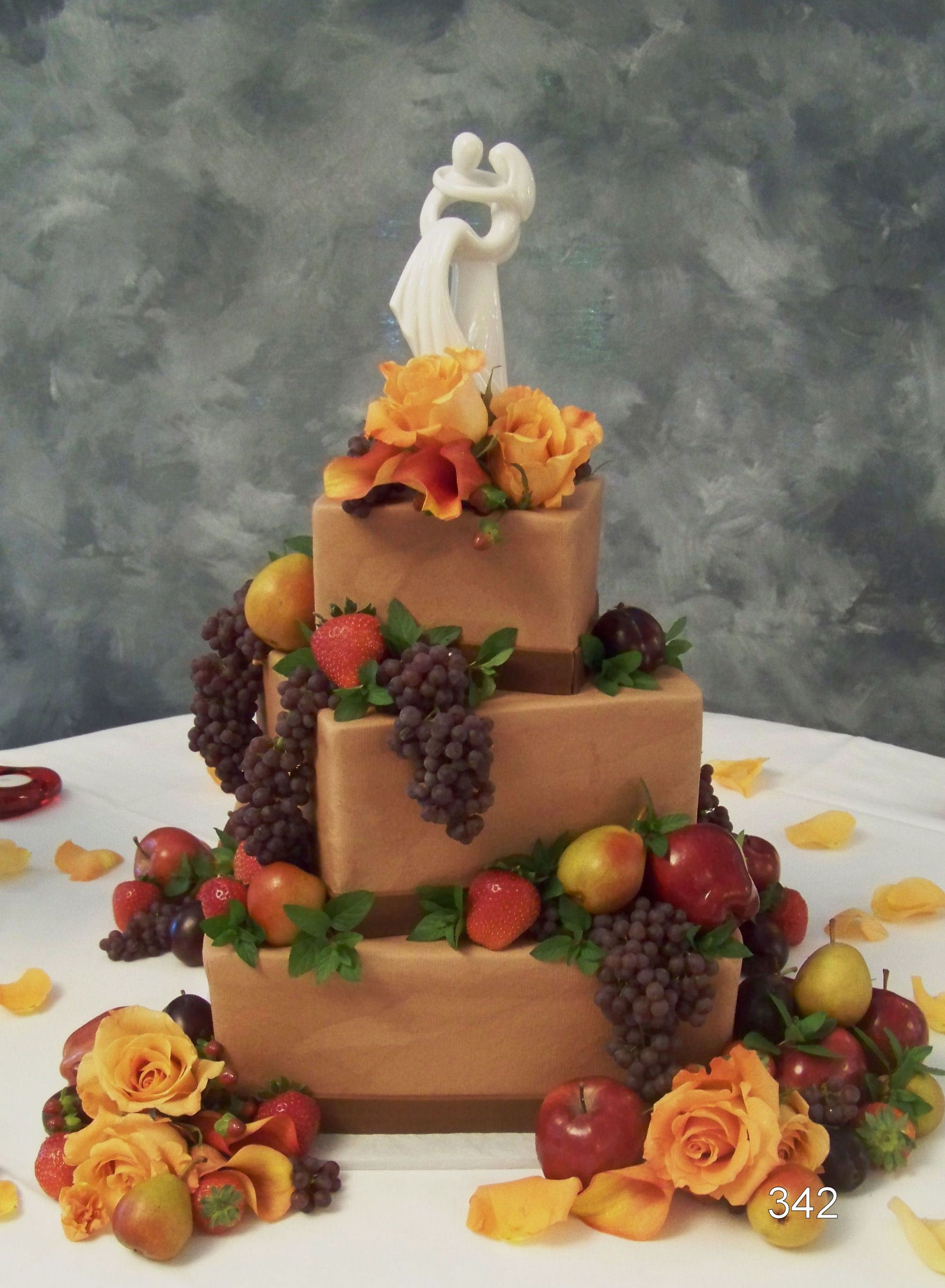 Three-tiered square wedding cake with fruit and flowers, topped with a bride and groom figurine