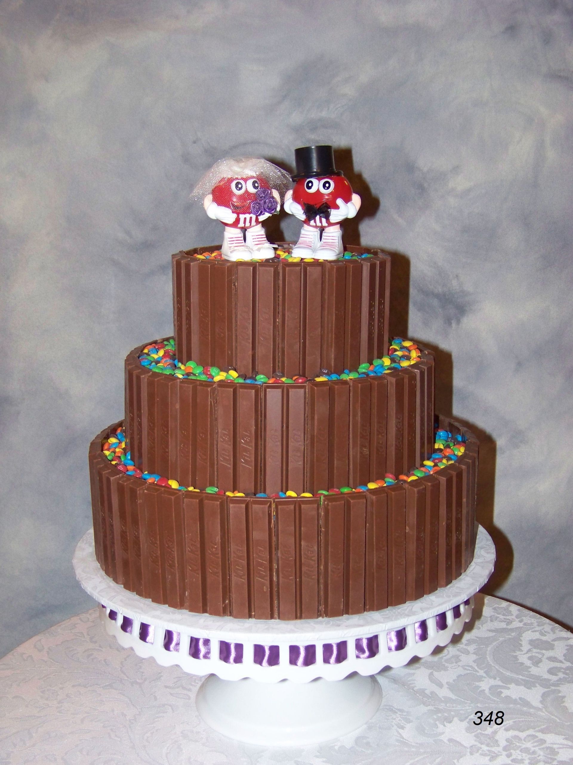 Three-tier chocolate-frosted cake with colorful sprinkles and two small figurines on top, on a white stand