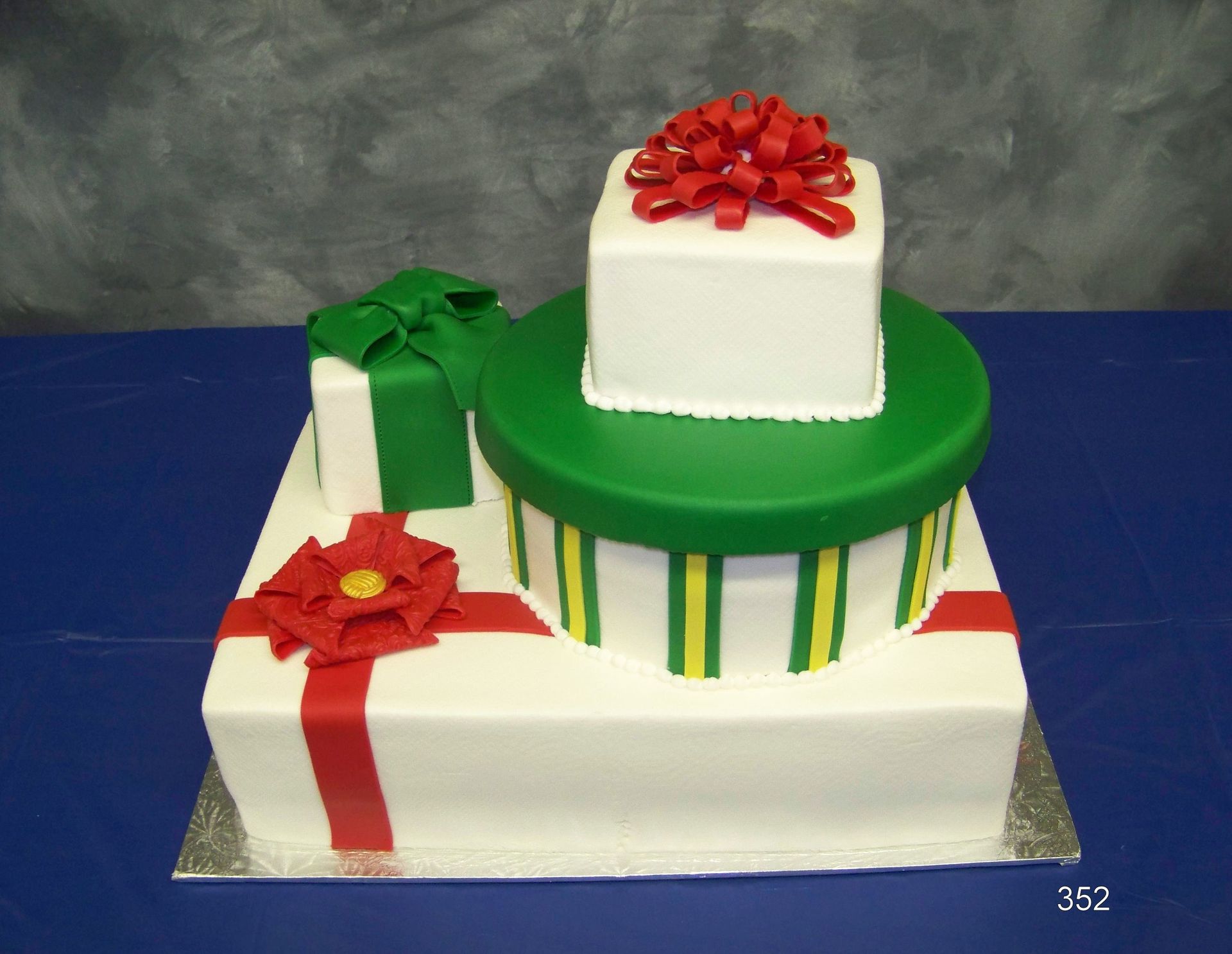 Two-tiered white gift cake with red bows and a green ribboned round topper on a blue surface.