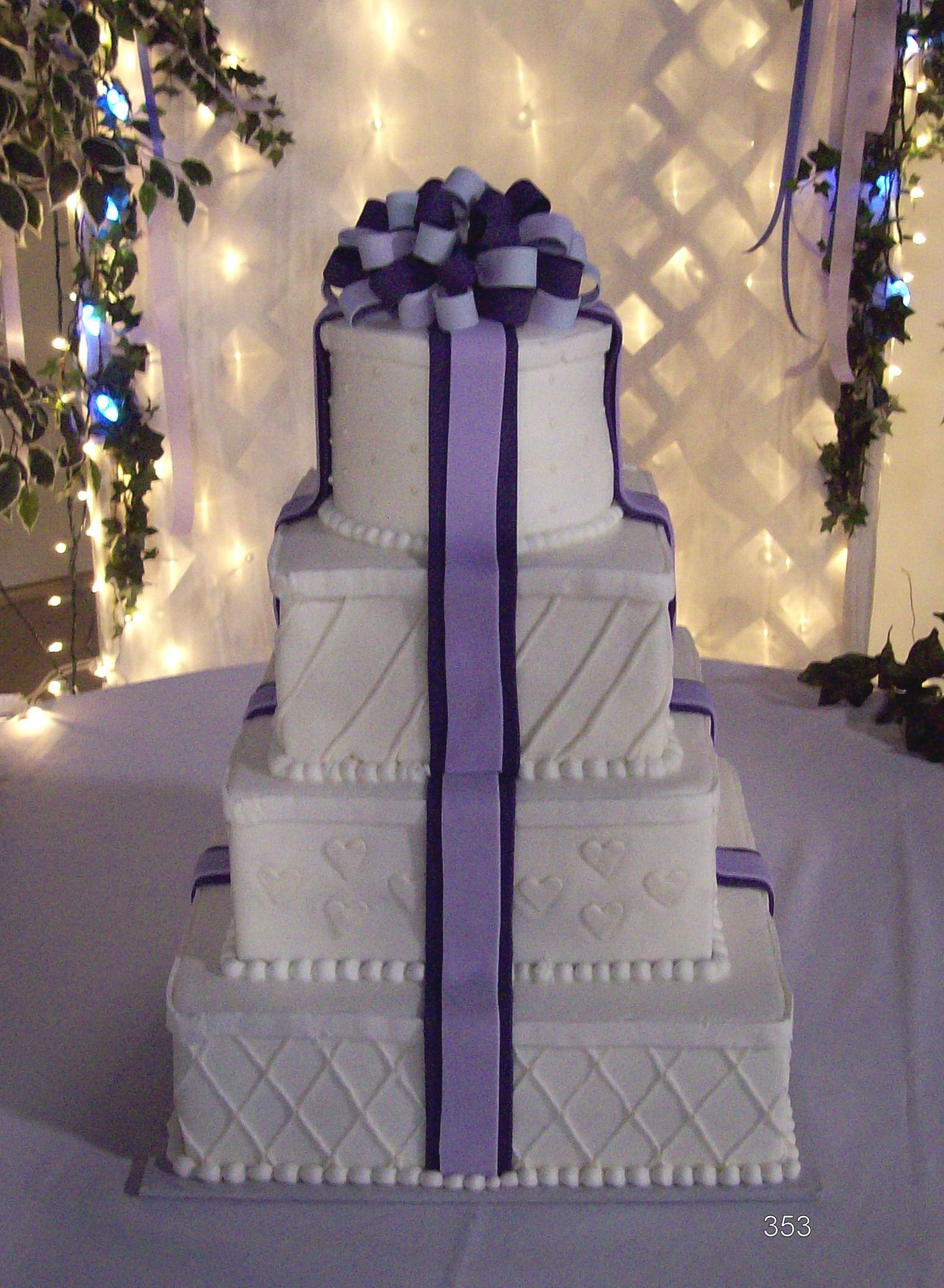 Four-tier white wedding cake with purple ribbon and bow, displayed on a decorated table.