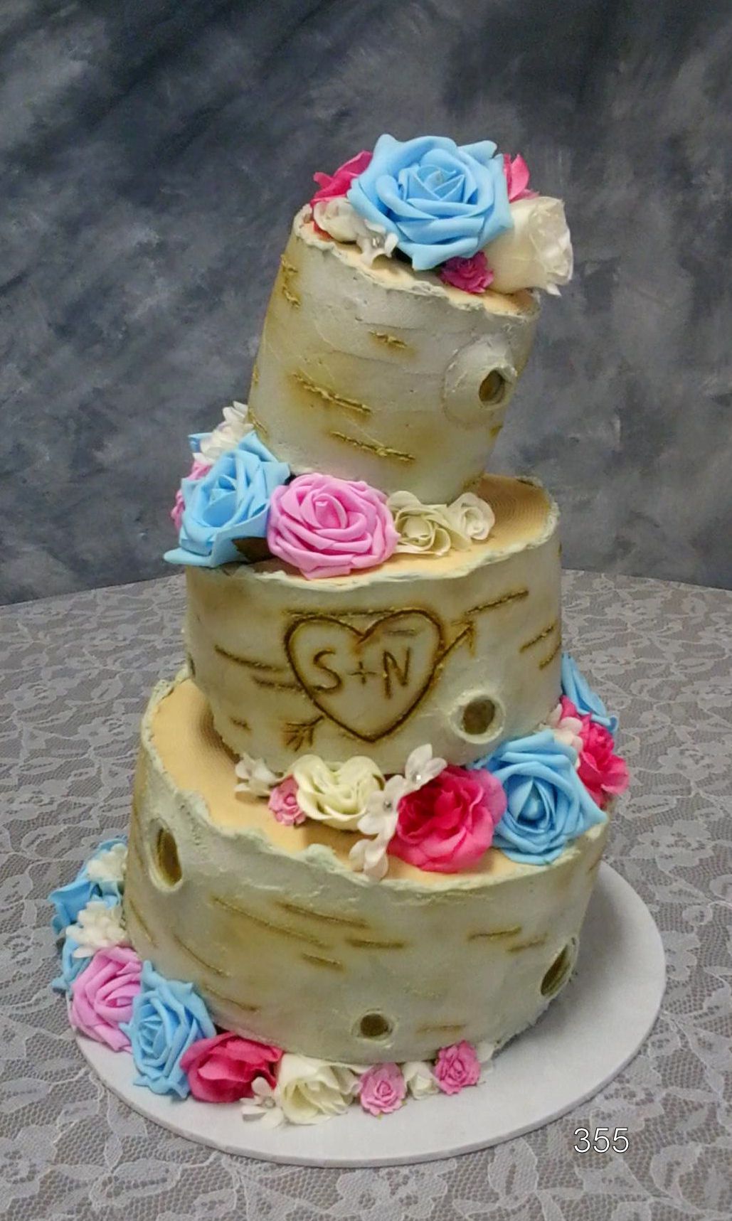 Three-tier rustic cake with gold accents and blue, pink, and white rosettes on a white stand