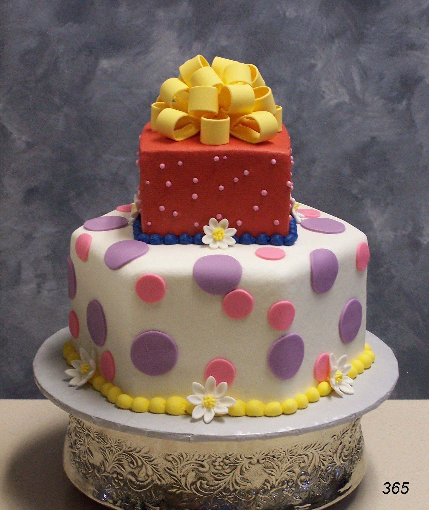 Two-tier decorated cake with white polka dots, red top tier, and yellow bow, on a silver cake stand