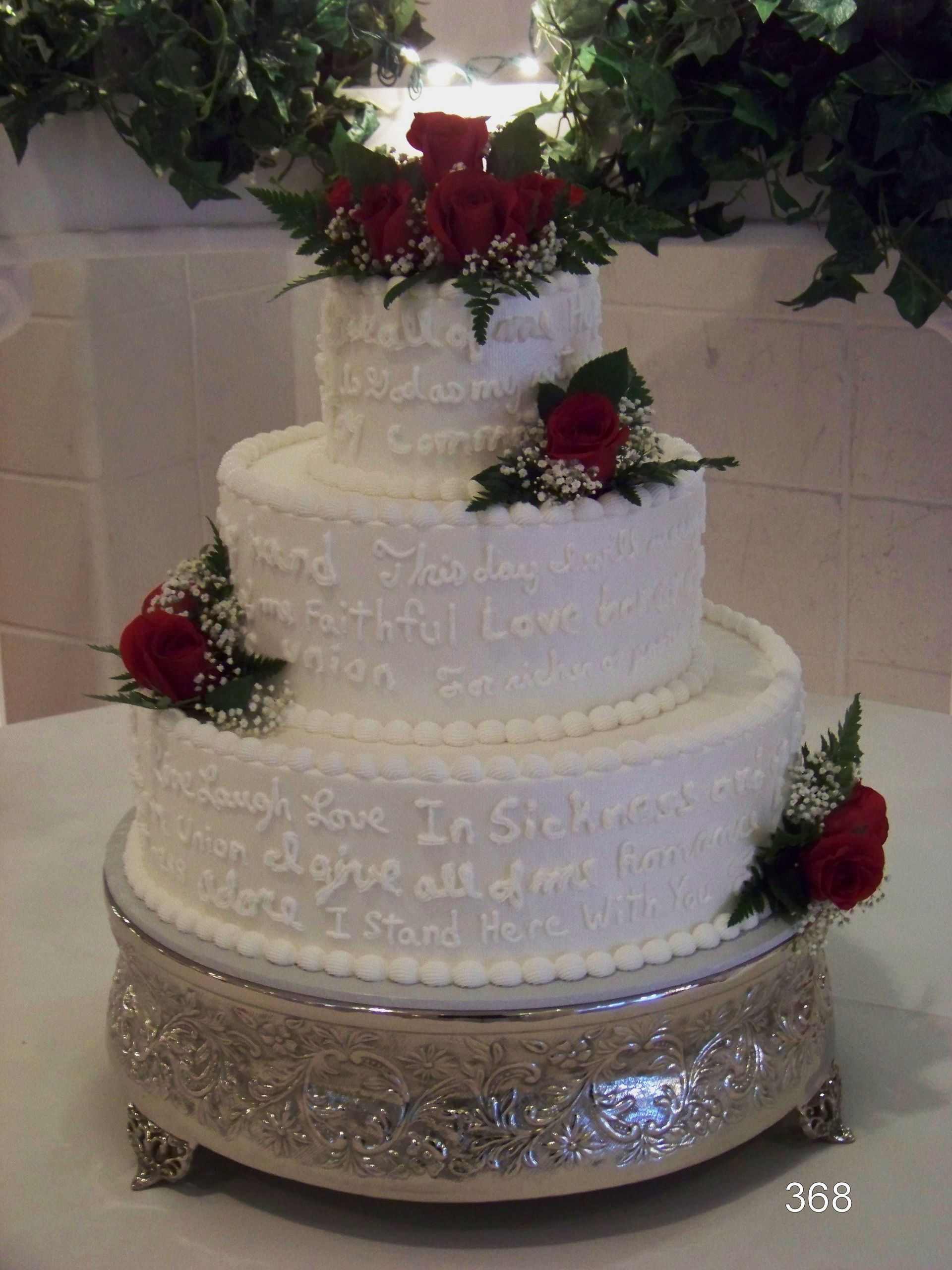 Three-tier white wedding cake with red roses and silver base stands on a decorated table.