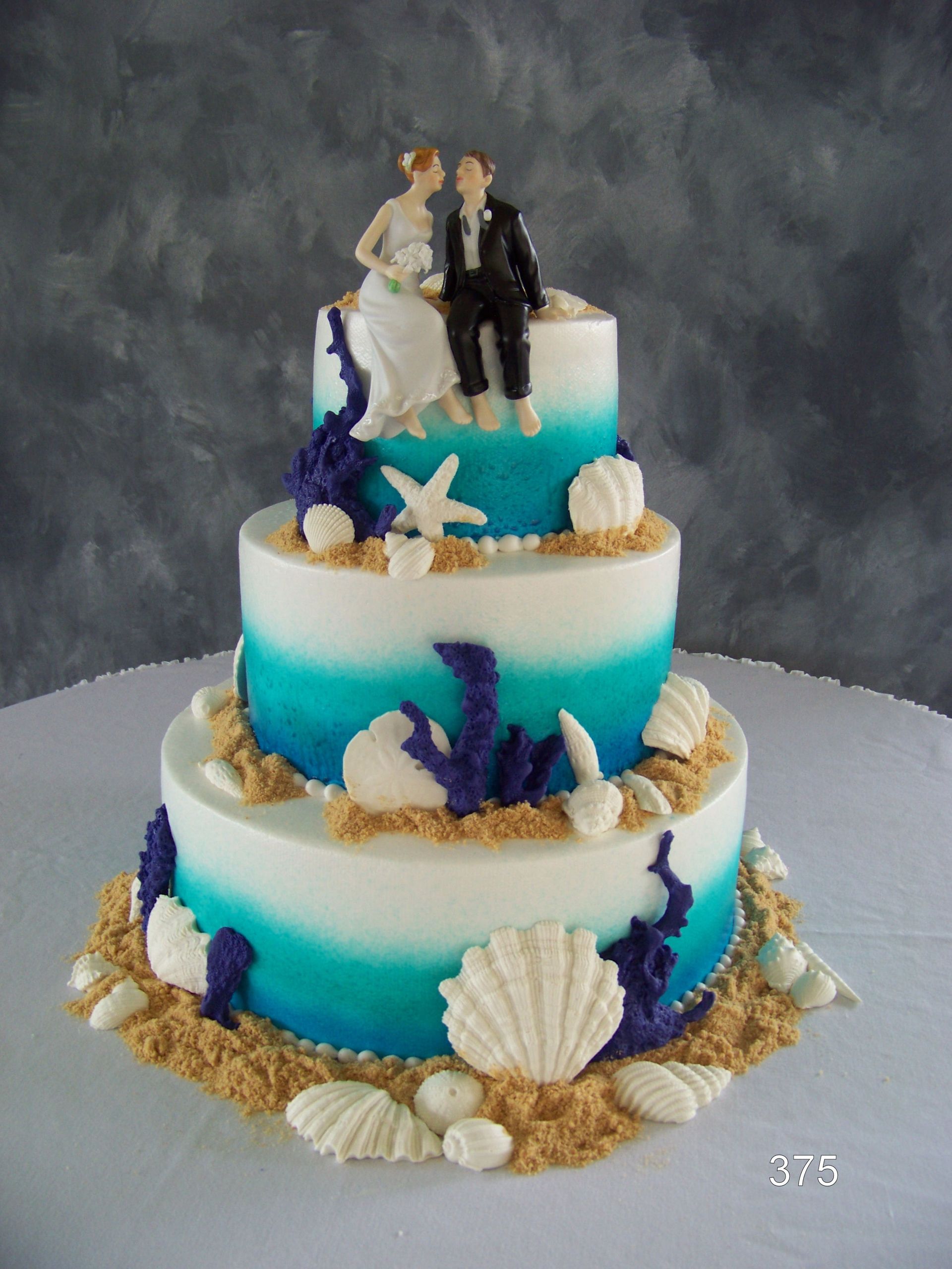 Three-tier blue-to-white ocean-themed cake with seashells, starfish, and a bride and groom topper