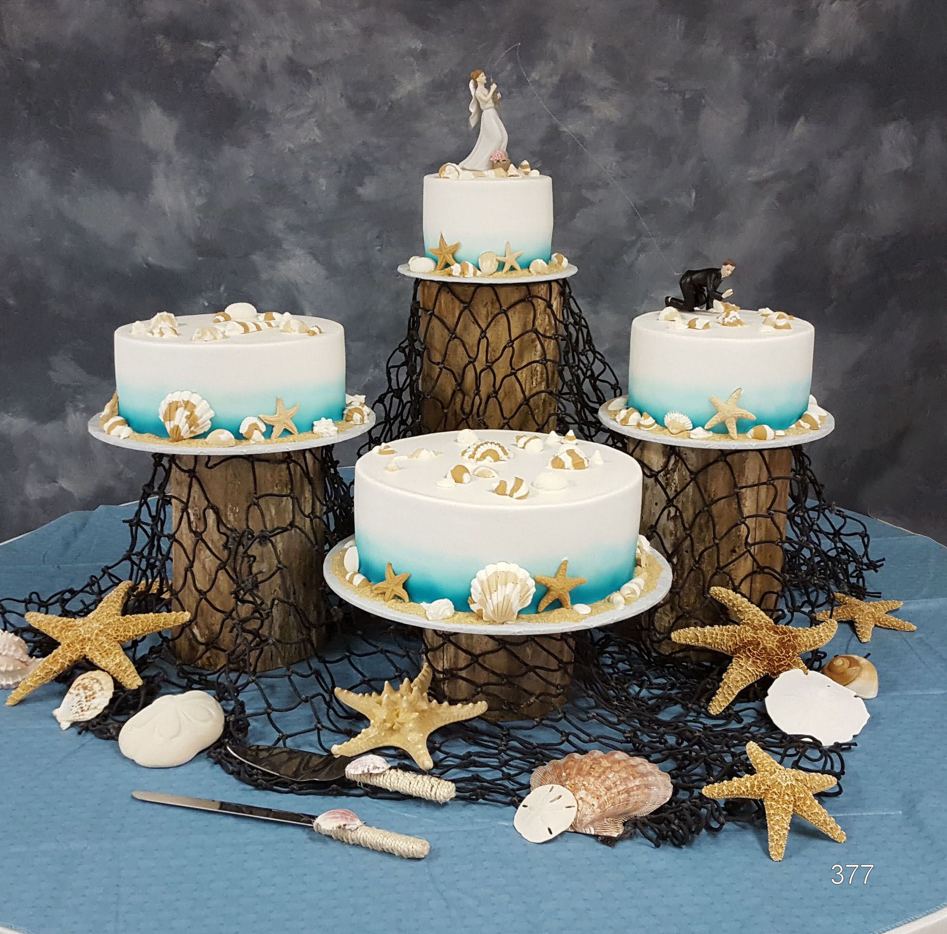 Four decorated white cakes on gold stands with seashell and starfish accents on a dark backdrop