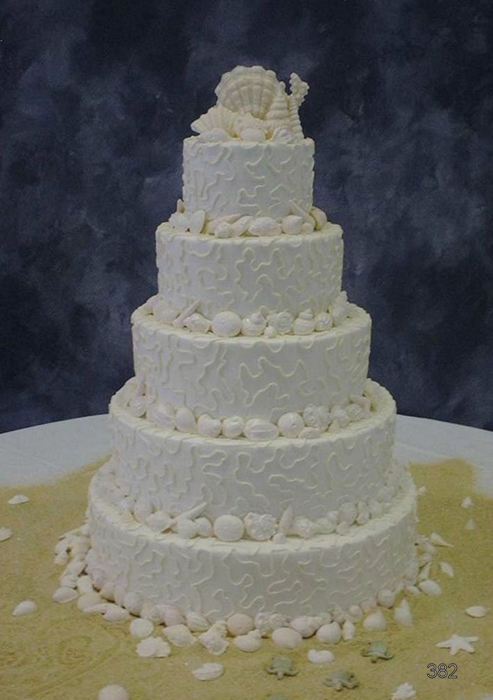 White tiered wedding cake with decorative frosting and white flowers on a table against a dark background