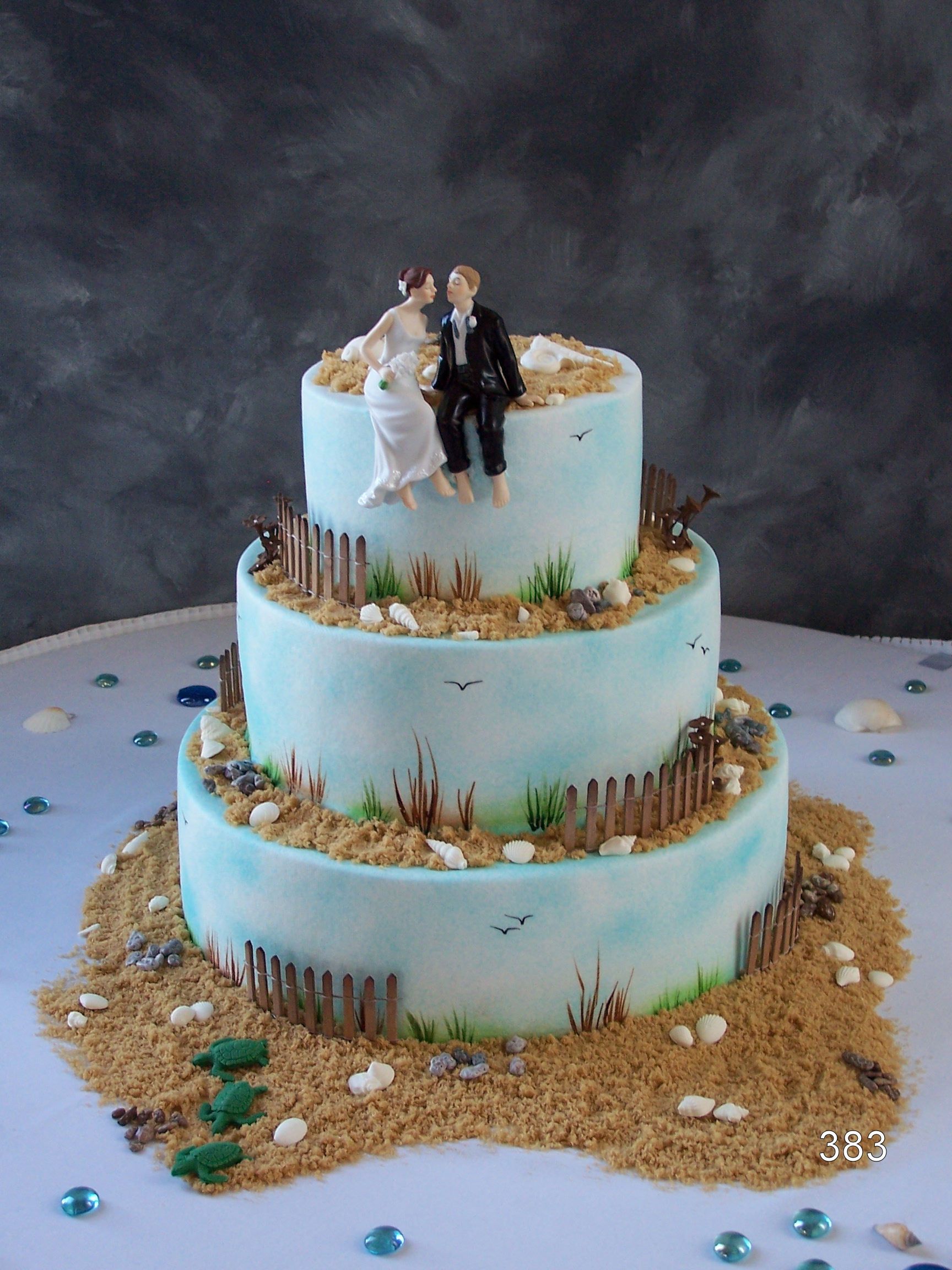 Three-tier blue wedding cake with bride and groom figurines on top and gold accents around the tiers