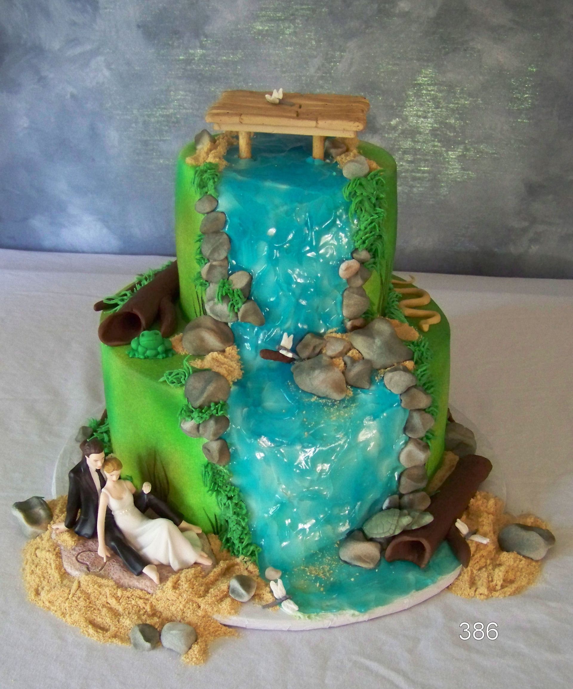 Cake decorated as a green hillside waterfall with blue stream, rocks, logs, and small animal figurines.