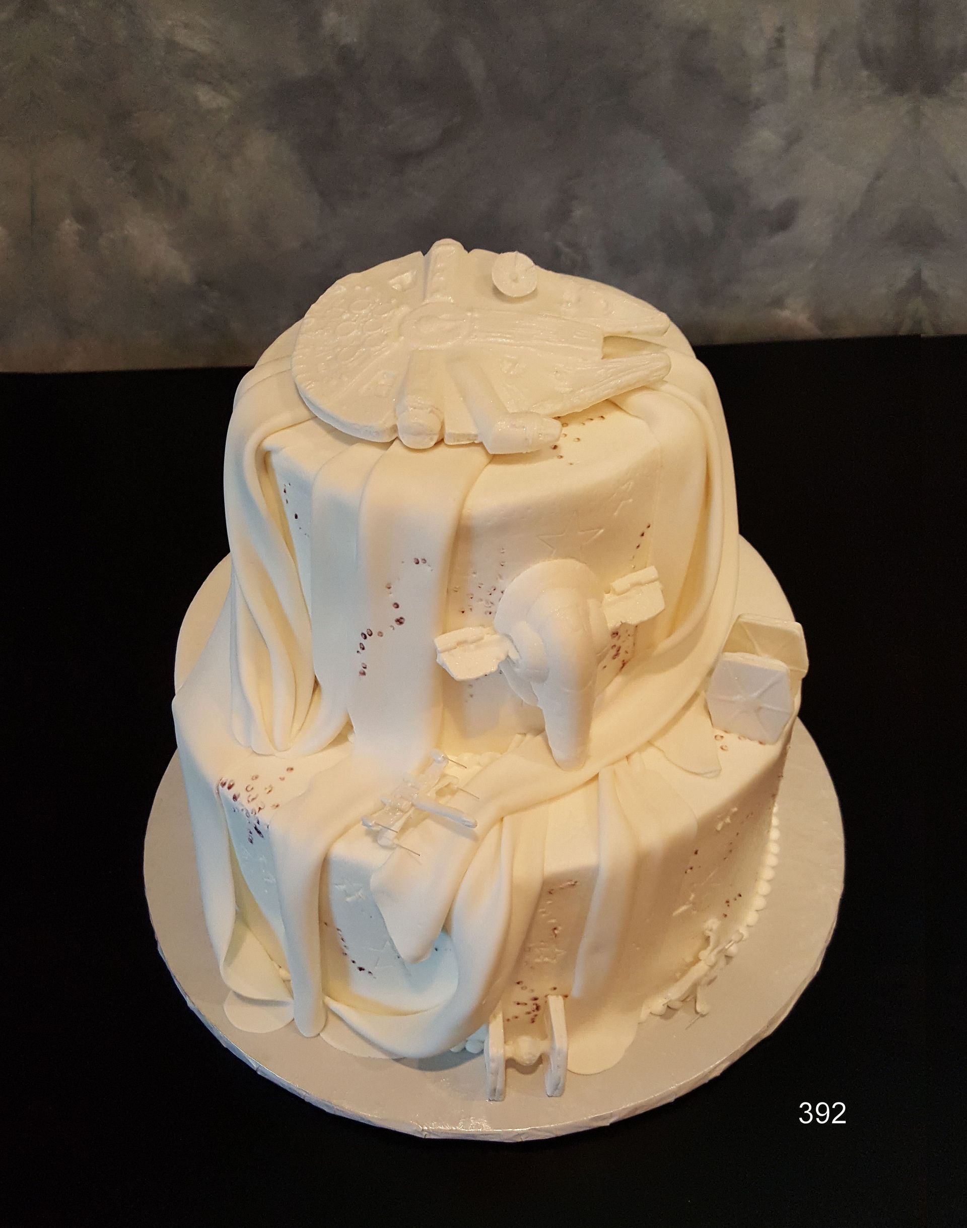 White frosted cake with draped icing and decorative top, displayed on a round board against a dark background