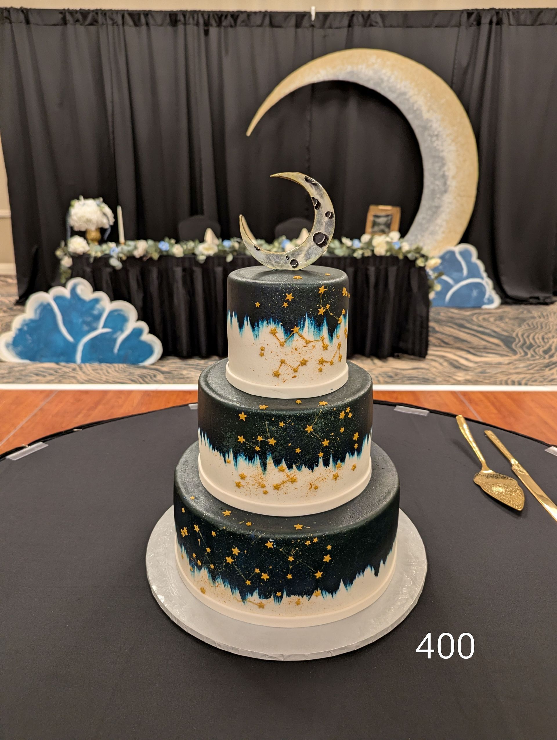 Three-tier navy and white cake with gold stars, set before a crescent moon backdrop and party dessert table