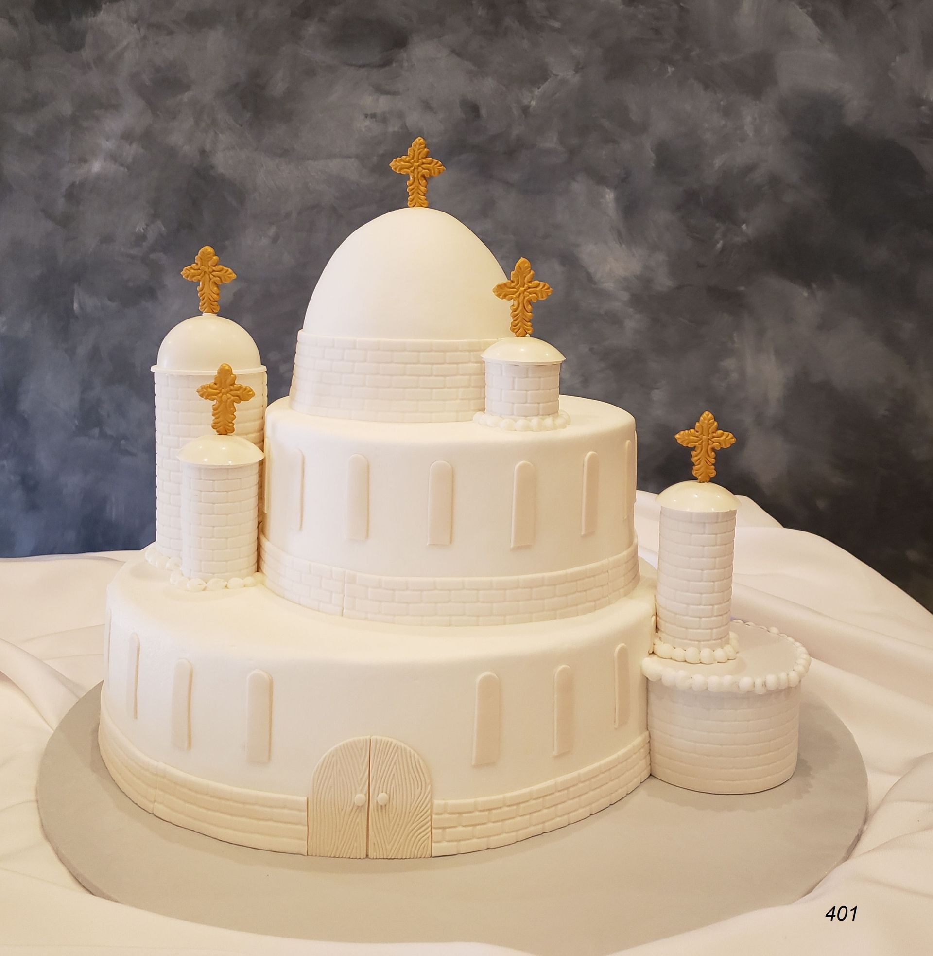 White tiered cake decorated like a church with gold crosses on a gray backdrop