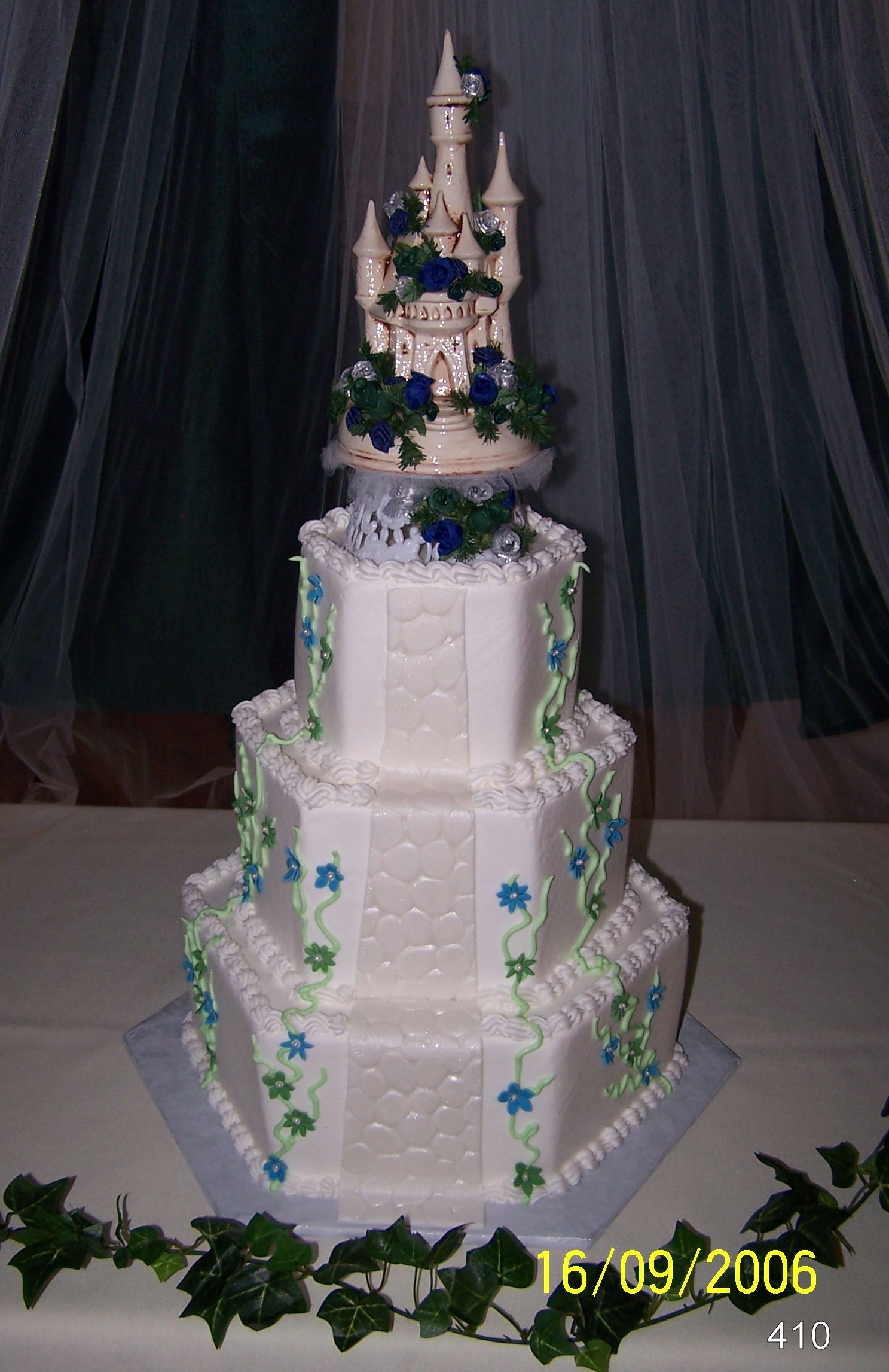 Tall white multi-tiered decorated cake with blue flowers and silver topper on a table, draped black backdrop