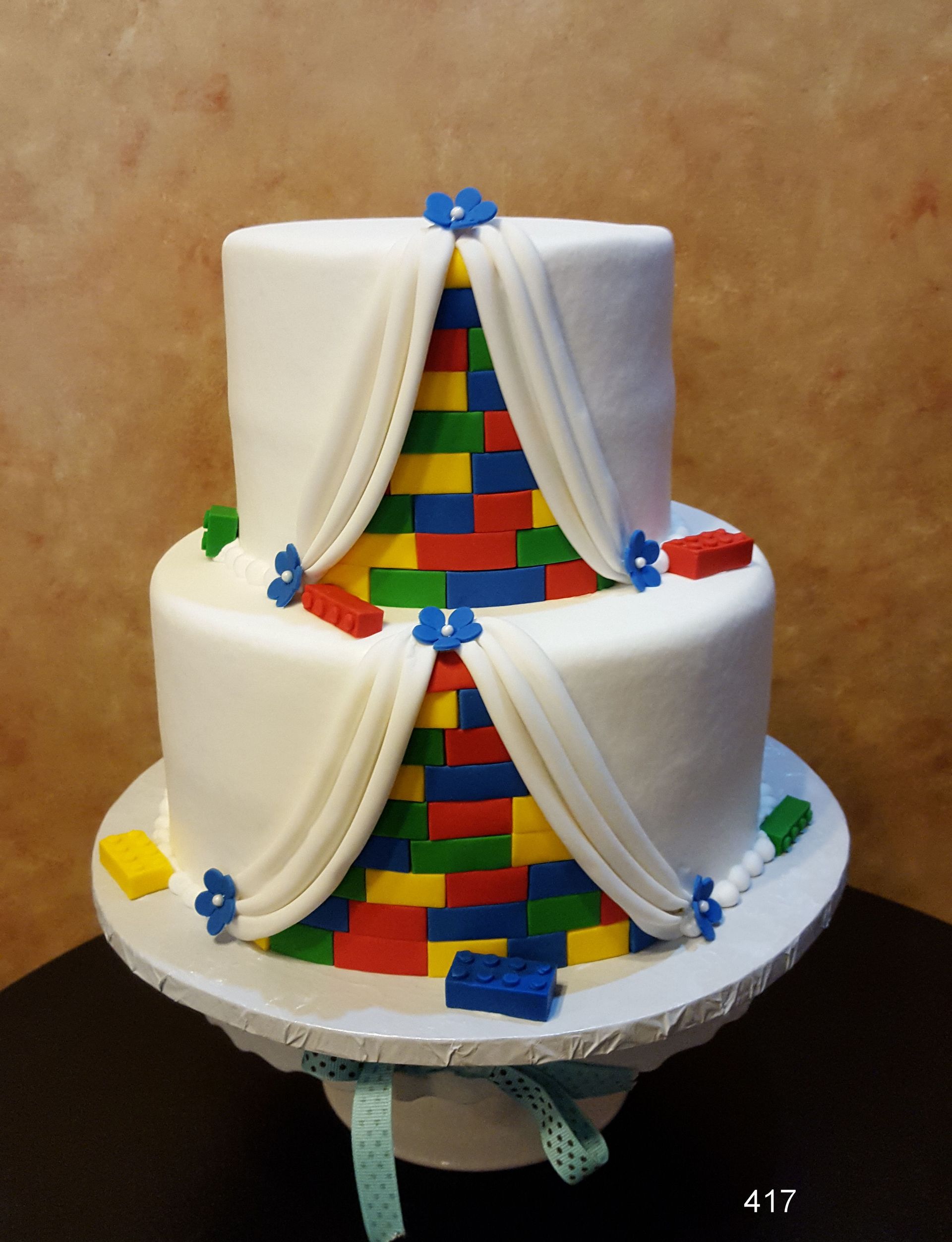 Two-tiered white cake with rainbow brick-pattern center and draped fondant decorations on a stand