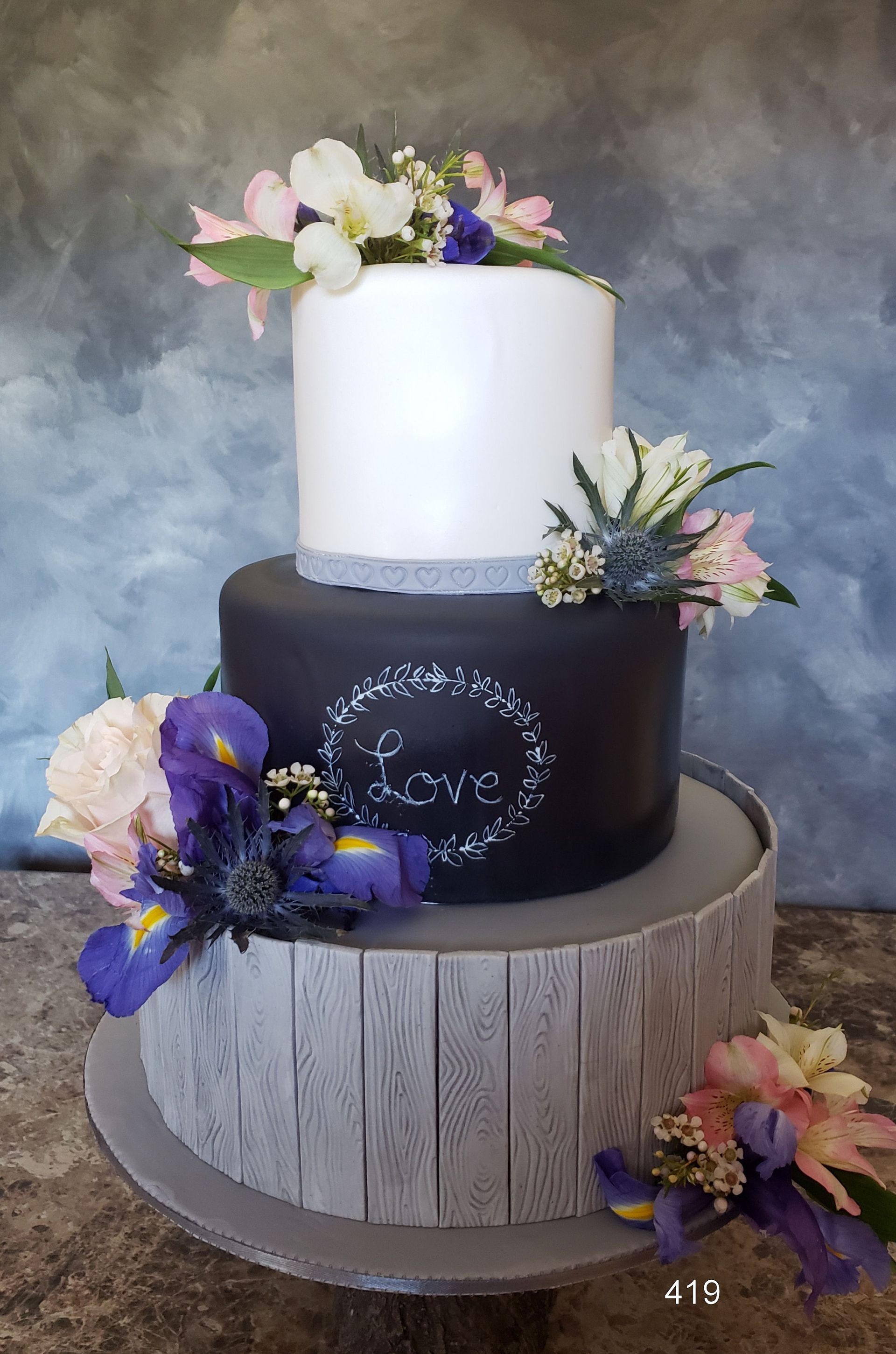 Three-tier black and white cake with purple and pink floral decorations on a rustic stand
