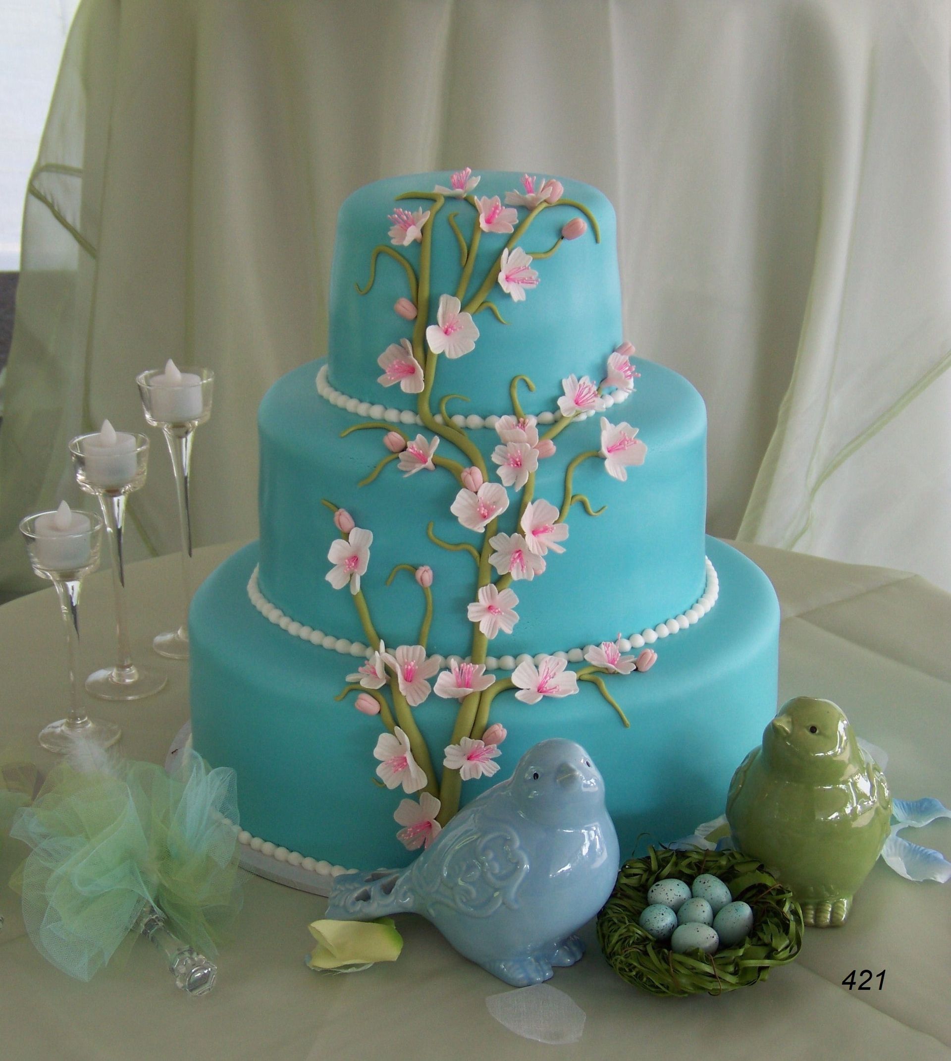 Turquoise three-tier cake with pink blossoms, flanked by ceramic birds and decorative glass flowers