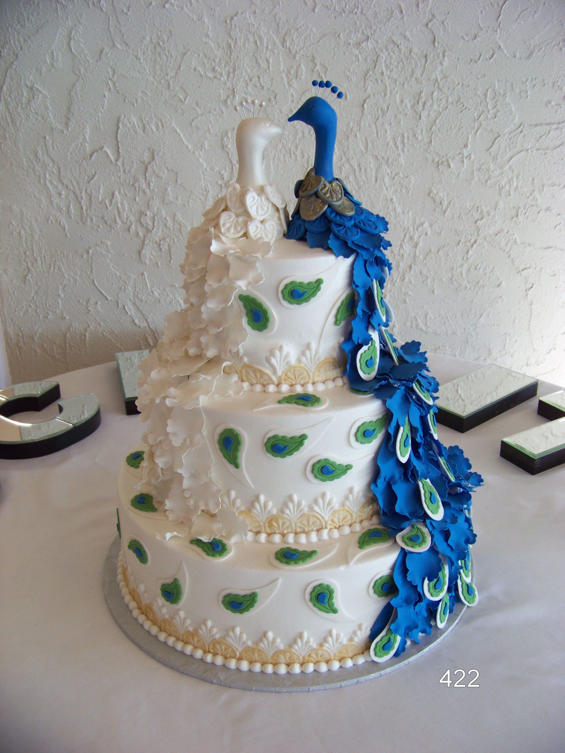 White tiered cake with green accents, white roses, and a blue peacock decoration cascading down the side