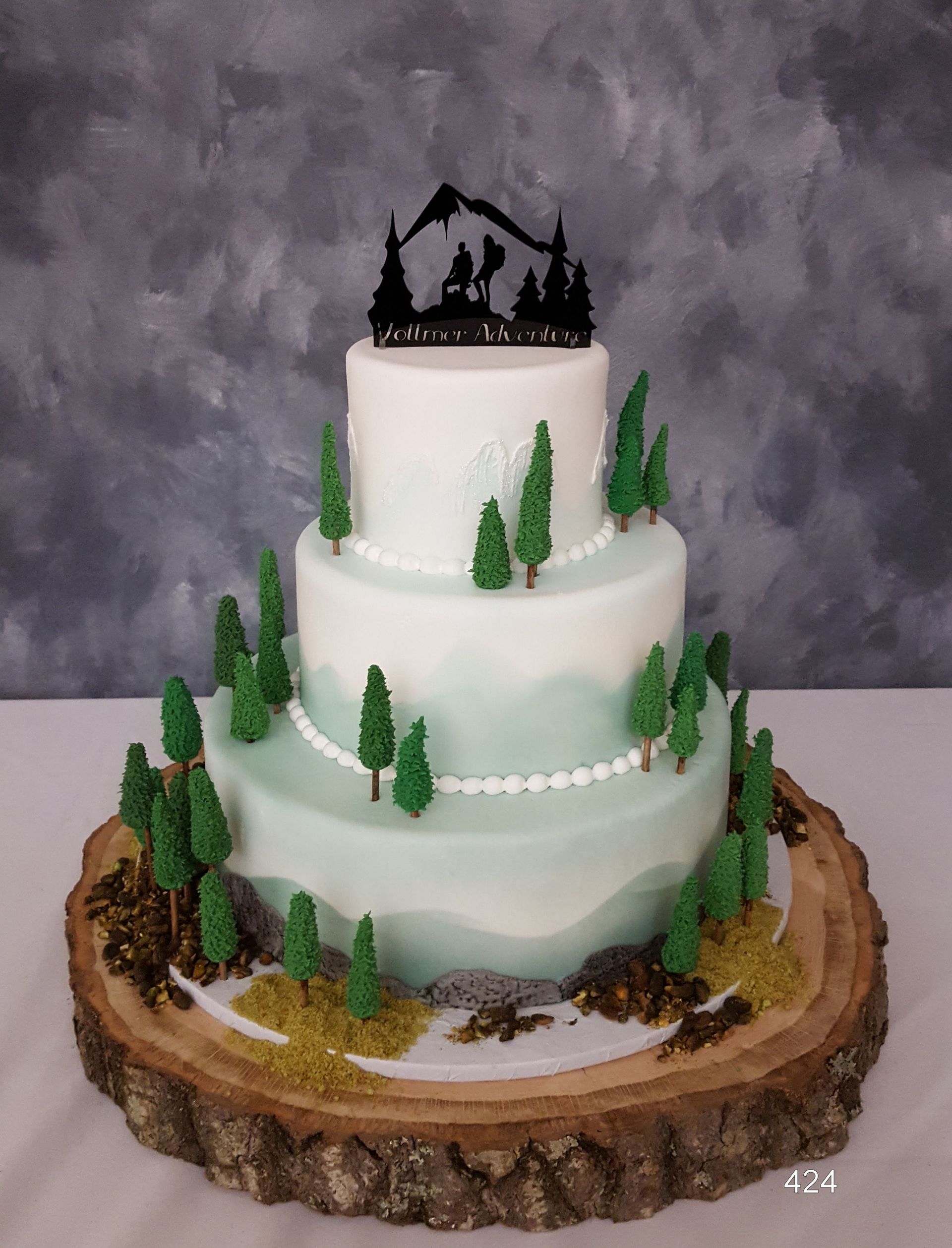 Three-tier white cake on a wood slab, decorated with evergreen trees and a black cabin topper.